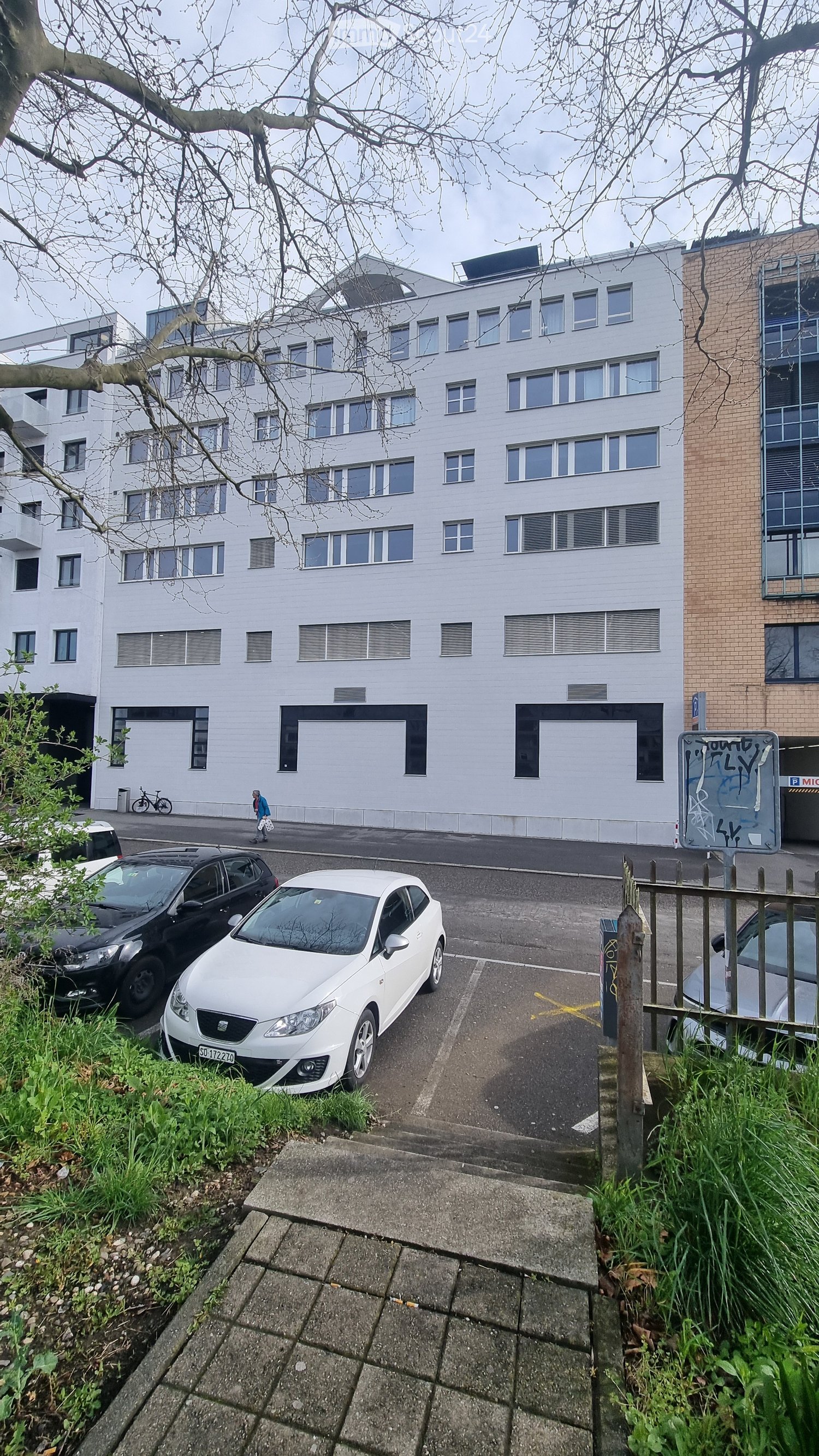 white 3 story building, windows, trees, sidewalk, cars parked, people walking, bicycles