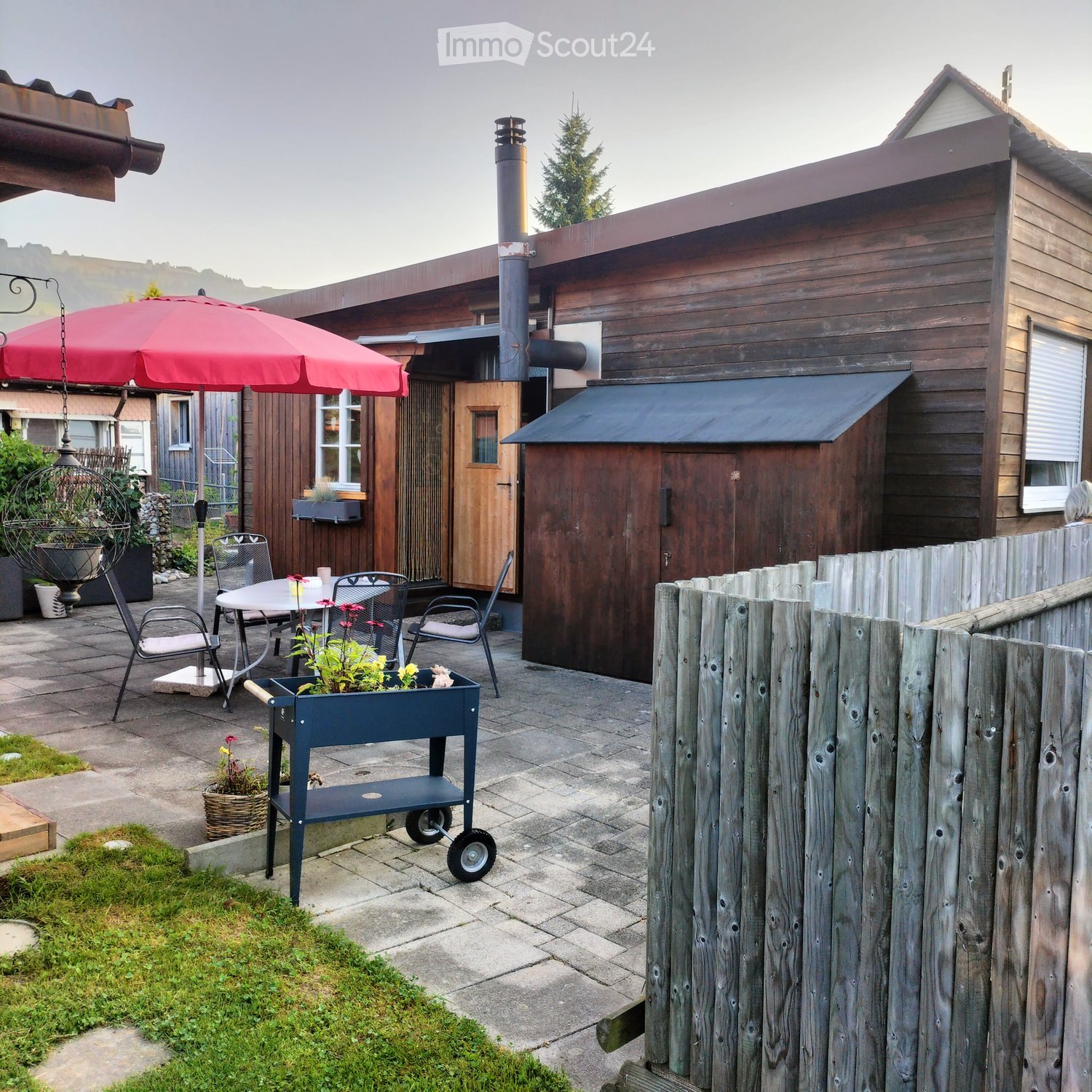 Brown wooden building, glass window, wooden patio furniture with red umbrella, wooden outdoor shed, brick-patterned flooring, grass lawn
