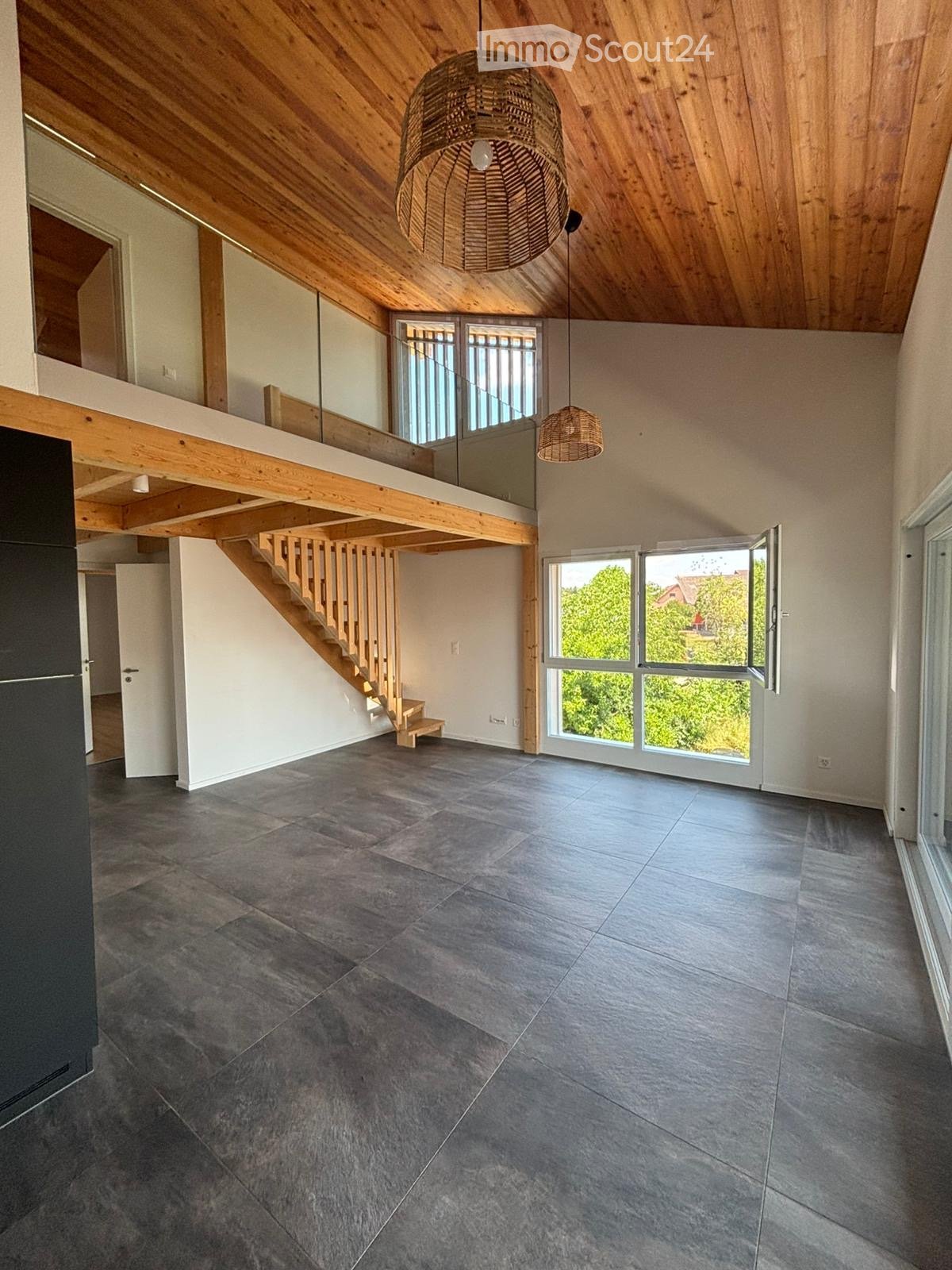 Open-plan living space with exposed wooden beams, grey tiled floor, large windows, and a wooden staircase leading to an upper level.