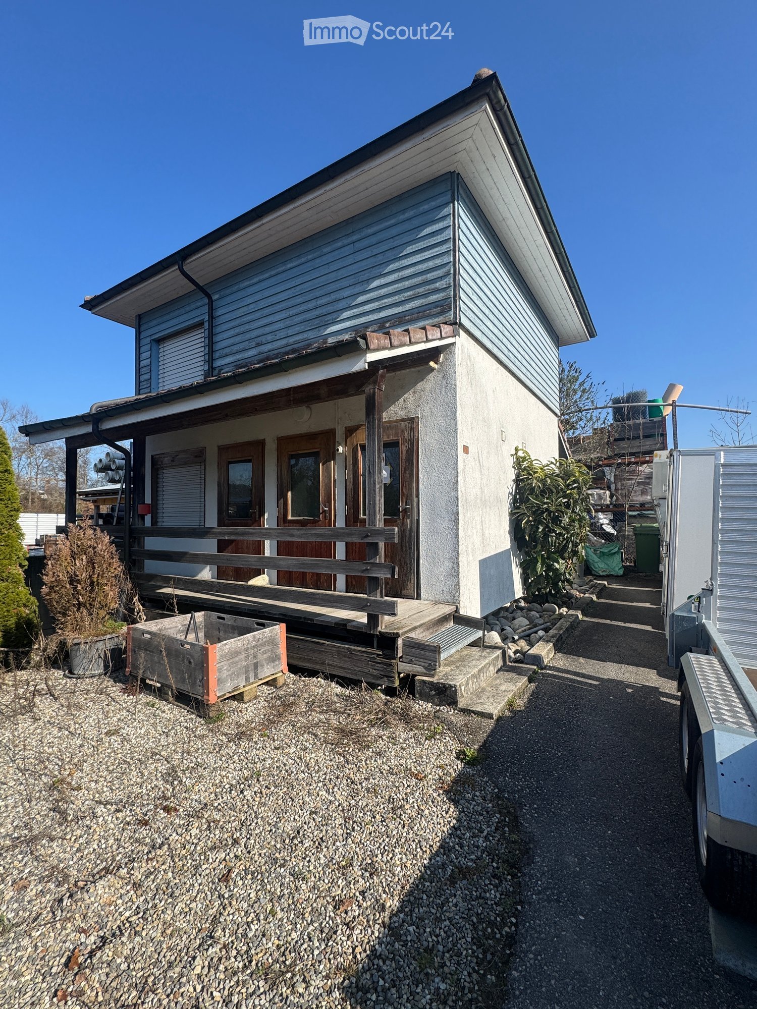 Two-story house, raised ground floor, wooden porch, large windows, blue siding, white exterior, gravel driveway