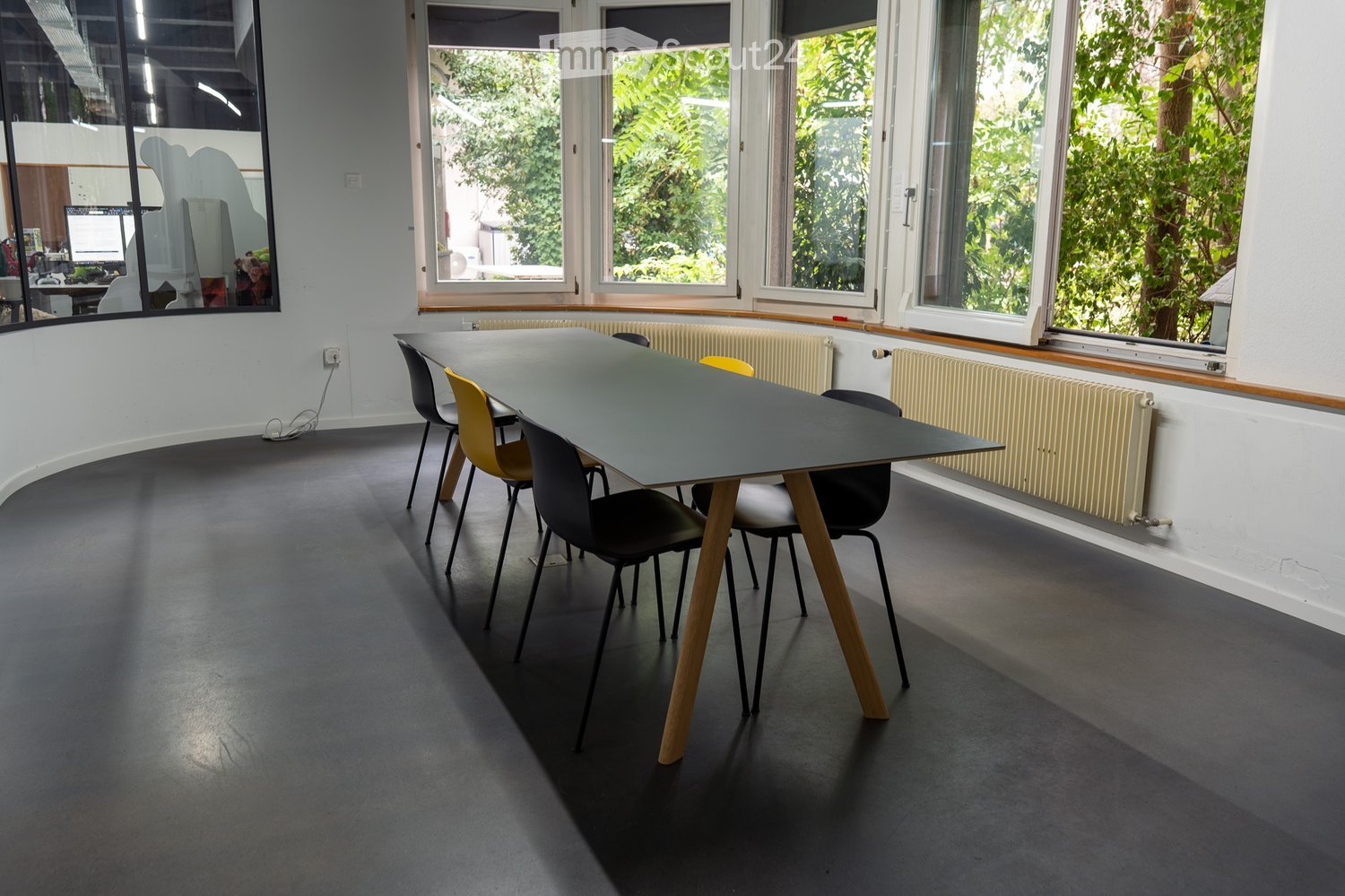 Modern, minimalist room with large windows, a grey table, and several chairs arranged around it. The floor is dark grey, and the walls are white.