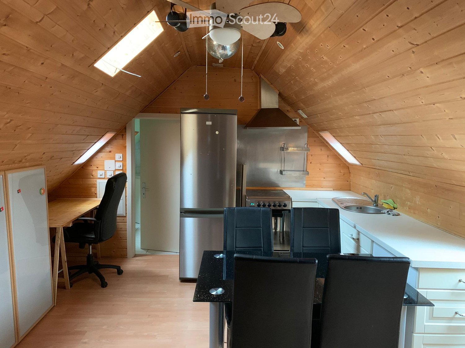Open kitchen with dining area and wooden walls and flooring, modern appliances, desk with chair, and a refrigerator.