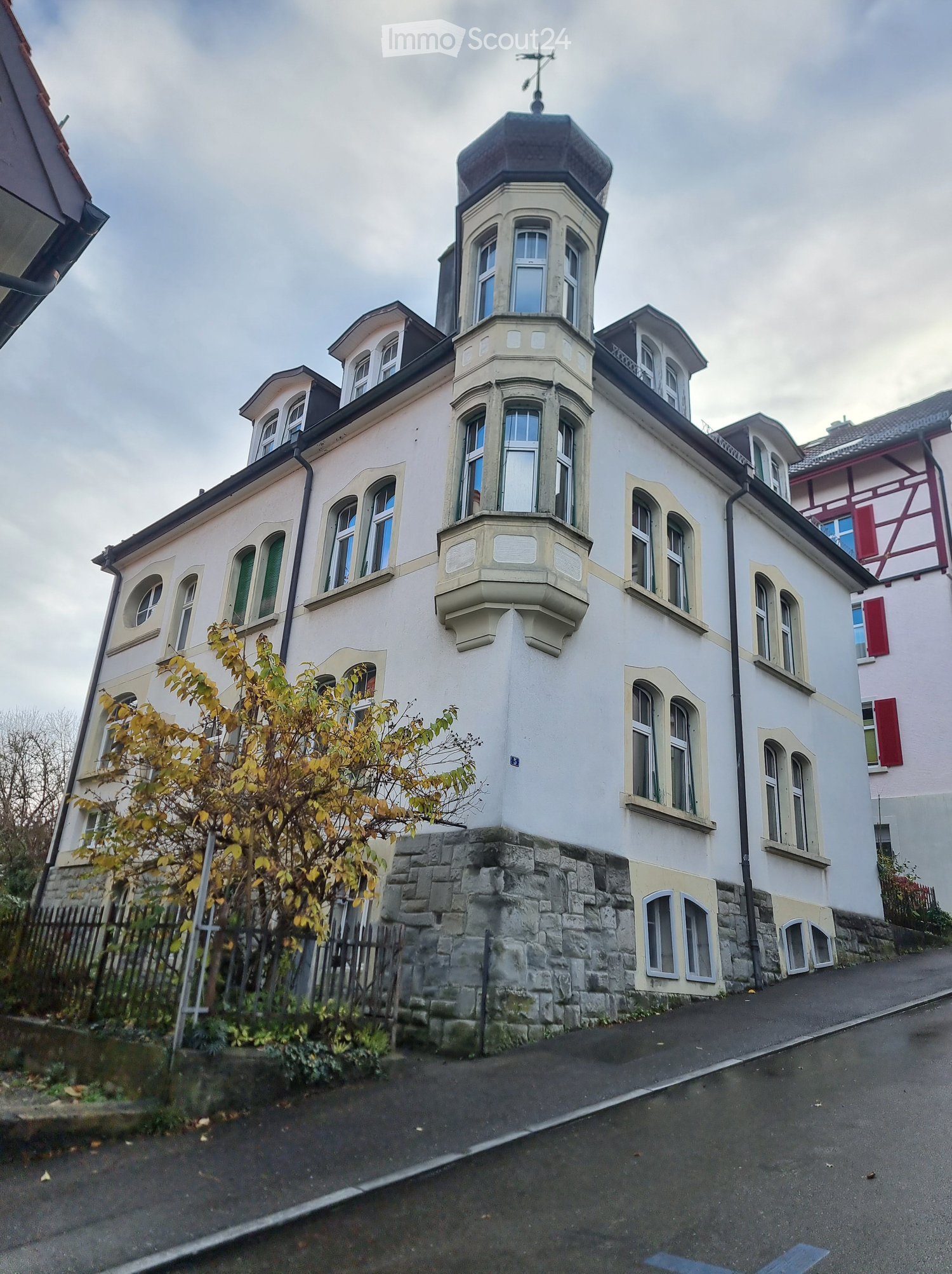 White building, multi-story, black roof, many arched windows, brick base, chimney, wrought iron fencing, green shutter on some windows