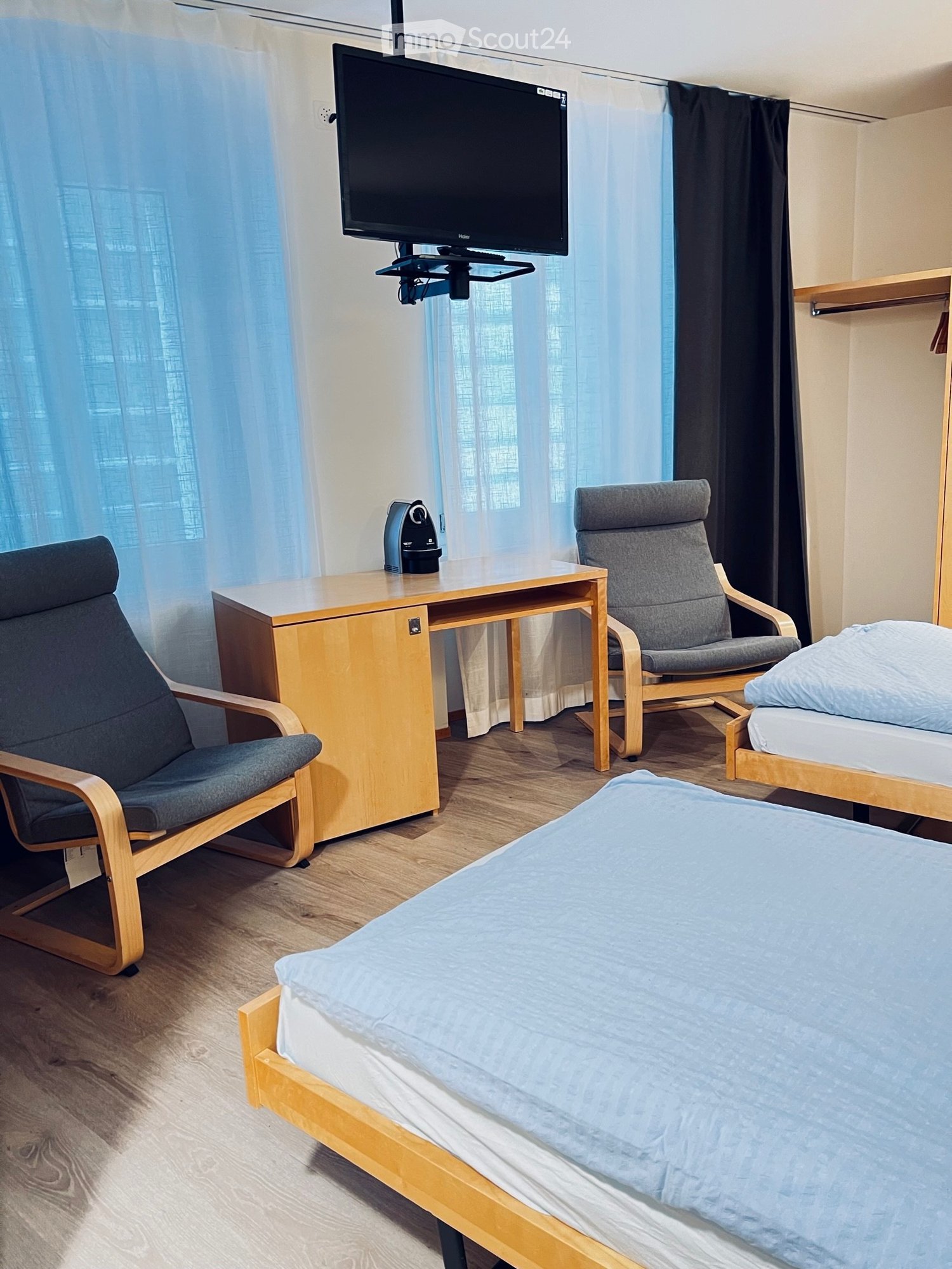 room with two beds, wooden desk with monitor, wooden chairs, floor lamp, shelf