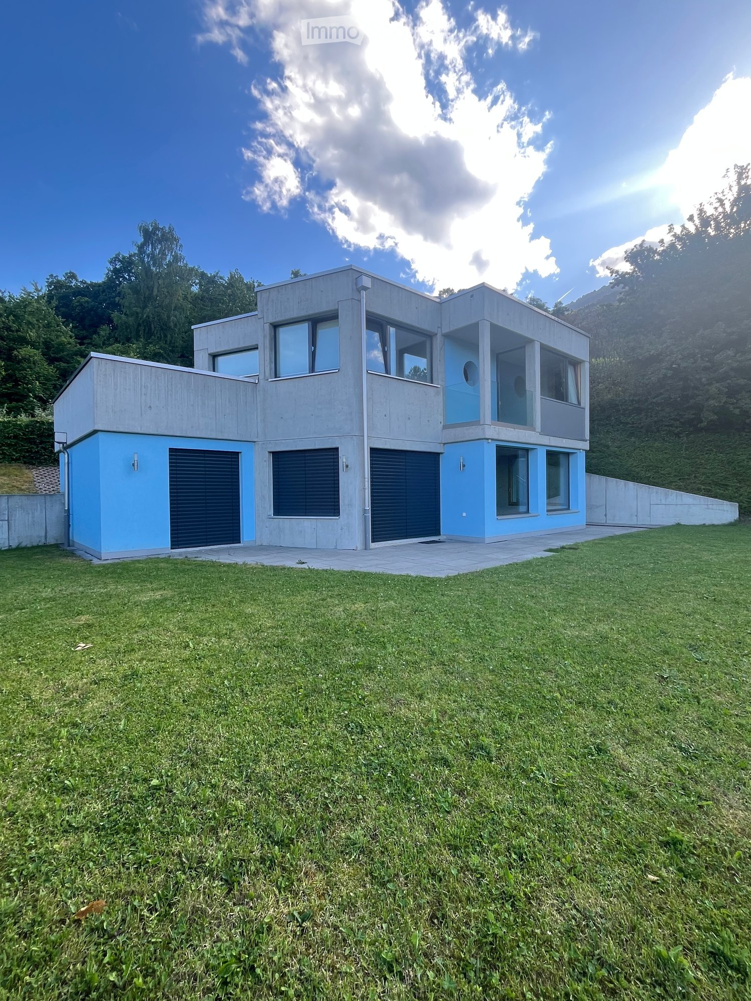 Modern two-story house with concrete and blue accents, large windows, and a spacious lawn in front.