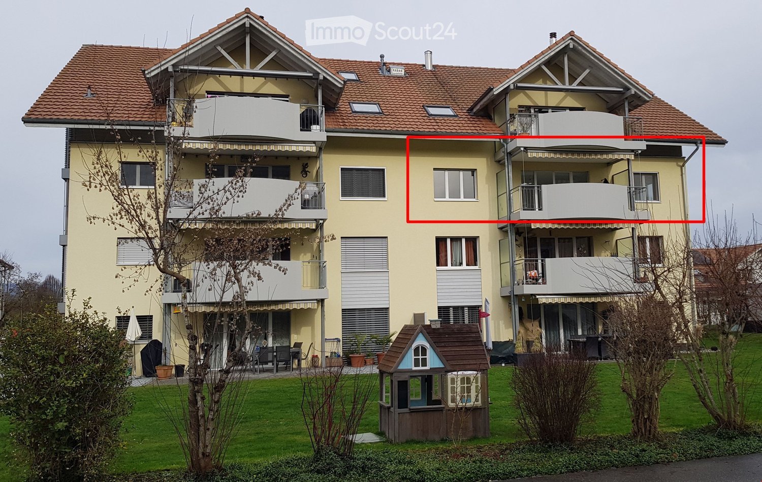 3 story apartment building, yellow exterior, red roof, balconies, fenced garden, large lawn