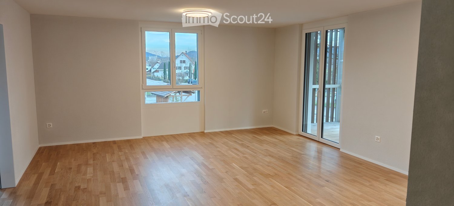 An empty room with hardwood floors, white walls, sliding doors leading to a balcony, and two windows that let in natural light and offer a view.