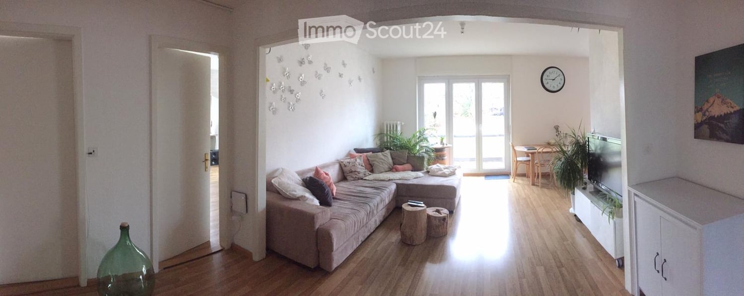 living room with wooden floor, white walls, large windows, L-shaped sofa, coffee table, TV, plants, clock, picture