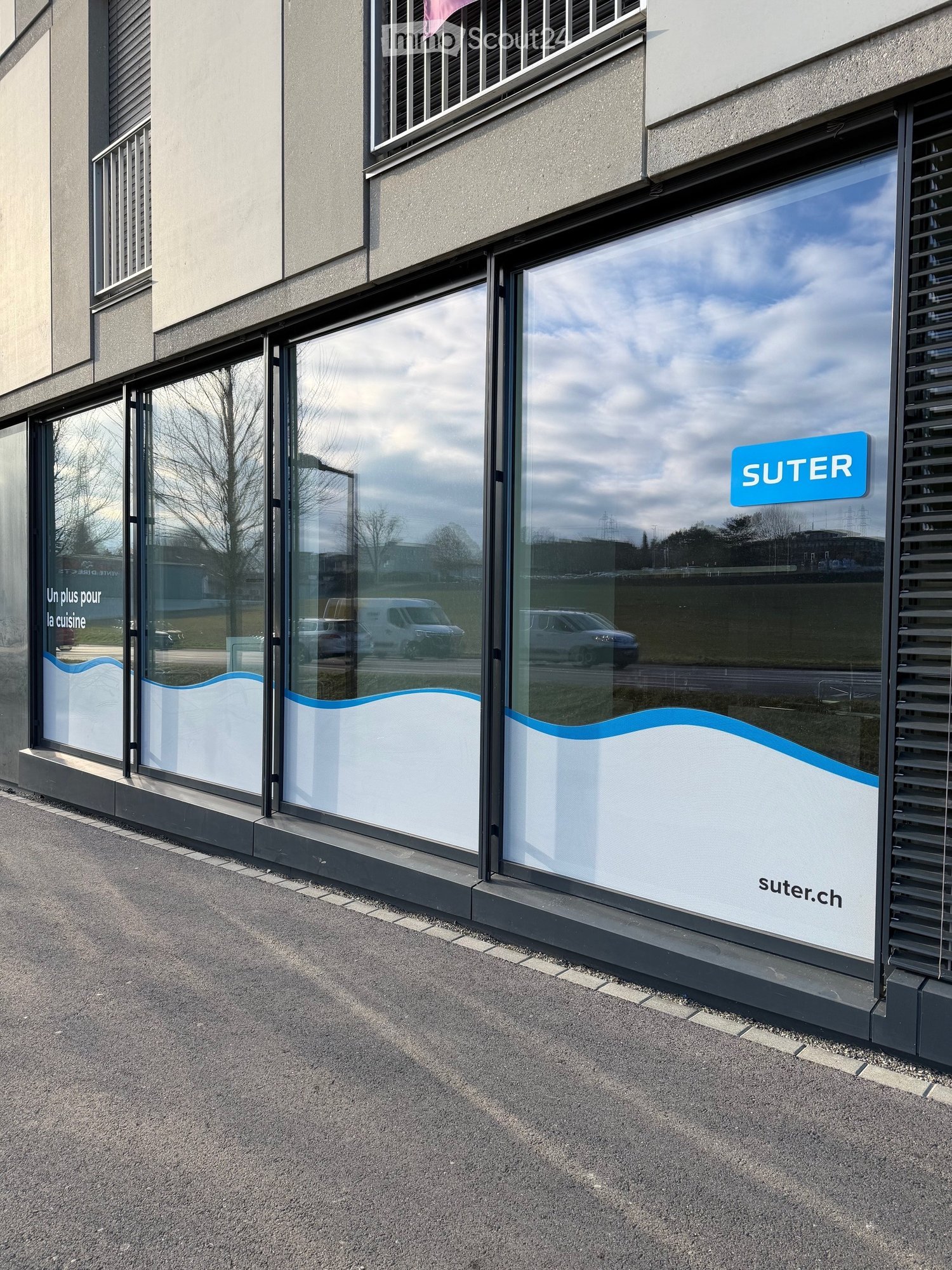 A commercial building with a glass window featuring 'SUTER' signage. The windows have wave-like patterns and French text 'Un plus pour la cuisine'. Reflections include cars and trees. The building has a concrete curb, and the ground is paved.