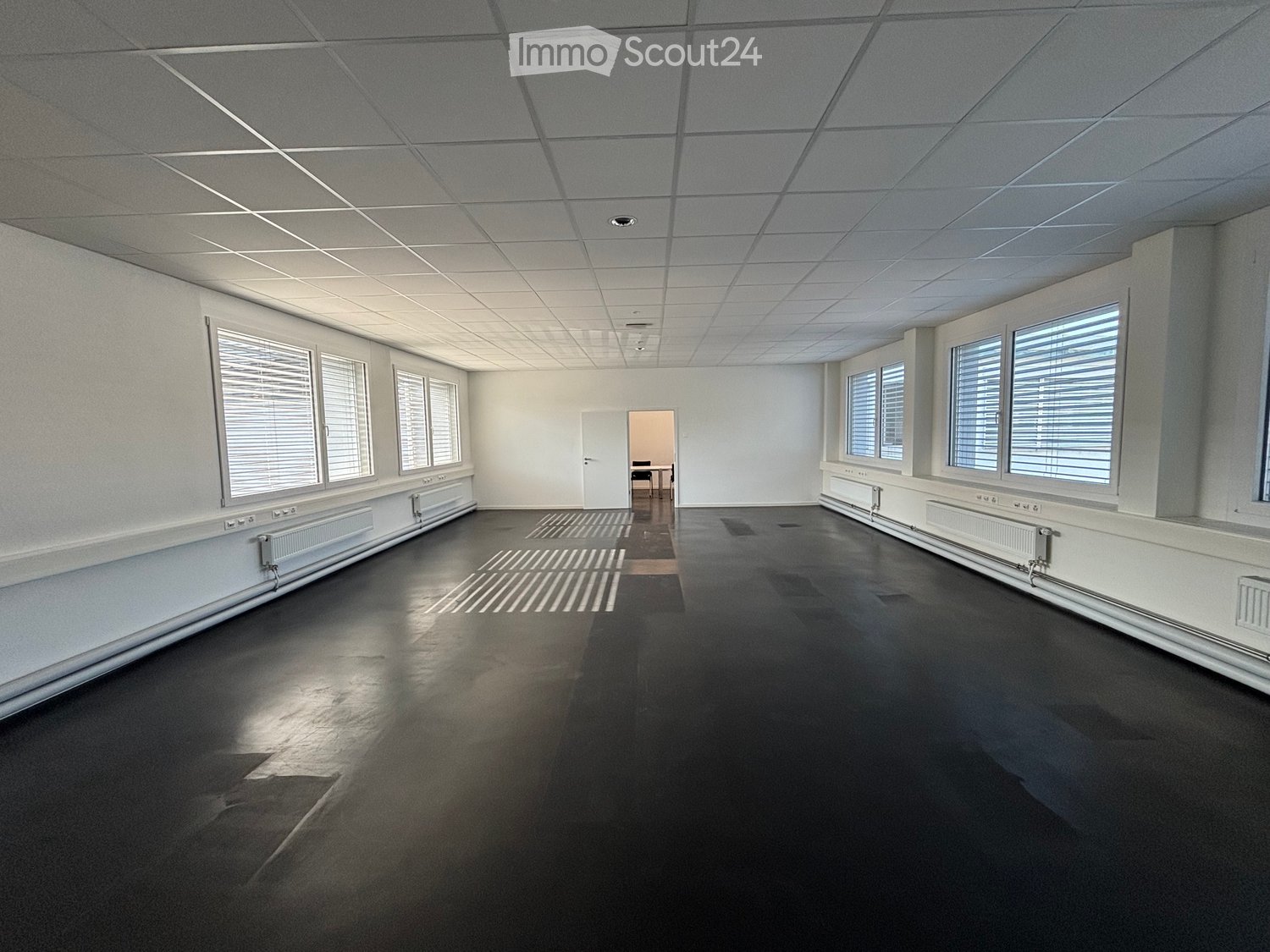 Empty room with white walls, black floor, large windows with blinds, and a doorway leading to another room.