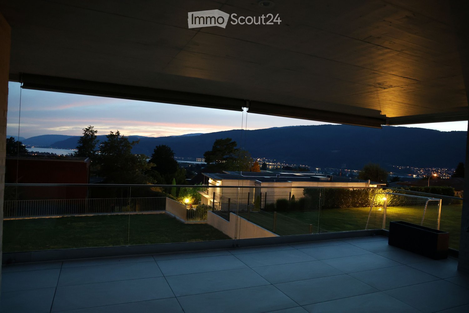 Balcony with tiled floor, glass railing, and a view of a cityscape and mountains at dusk.