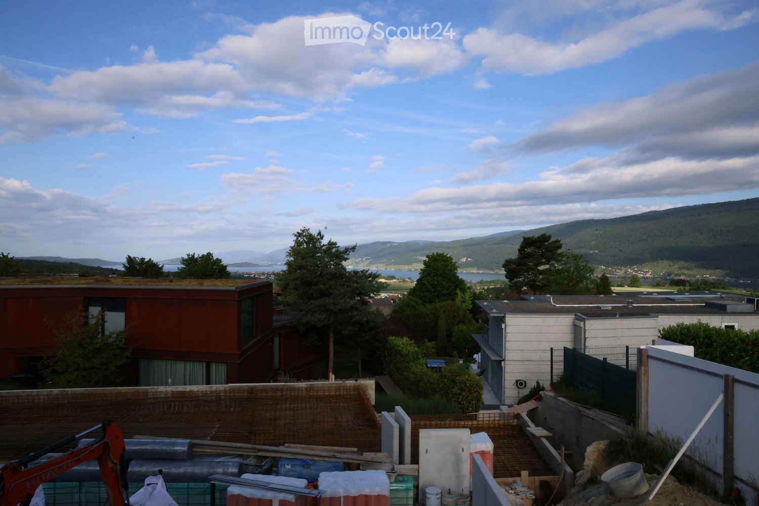 View of a modern residential area with contemporary buildings, greenery, and a scenic mountain and lake view in the background.