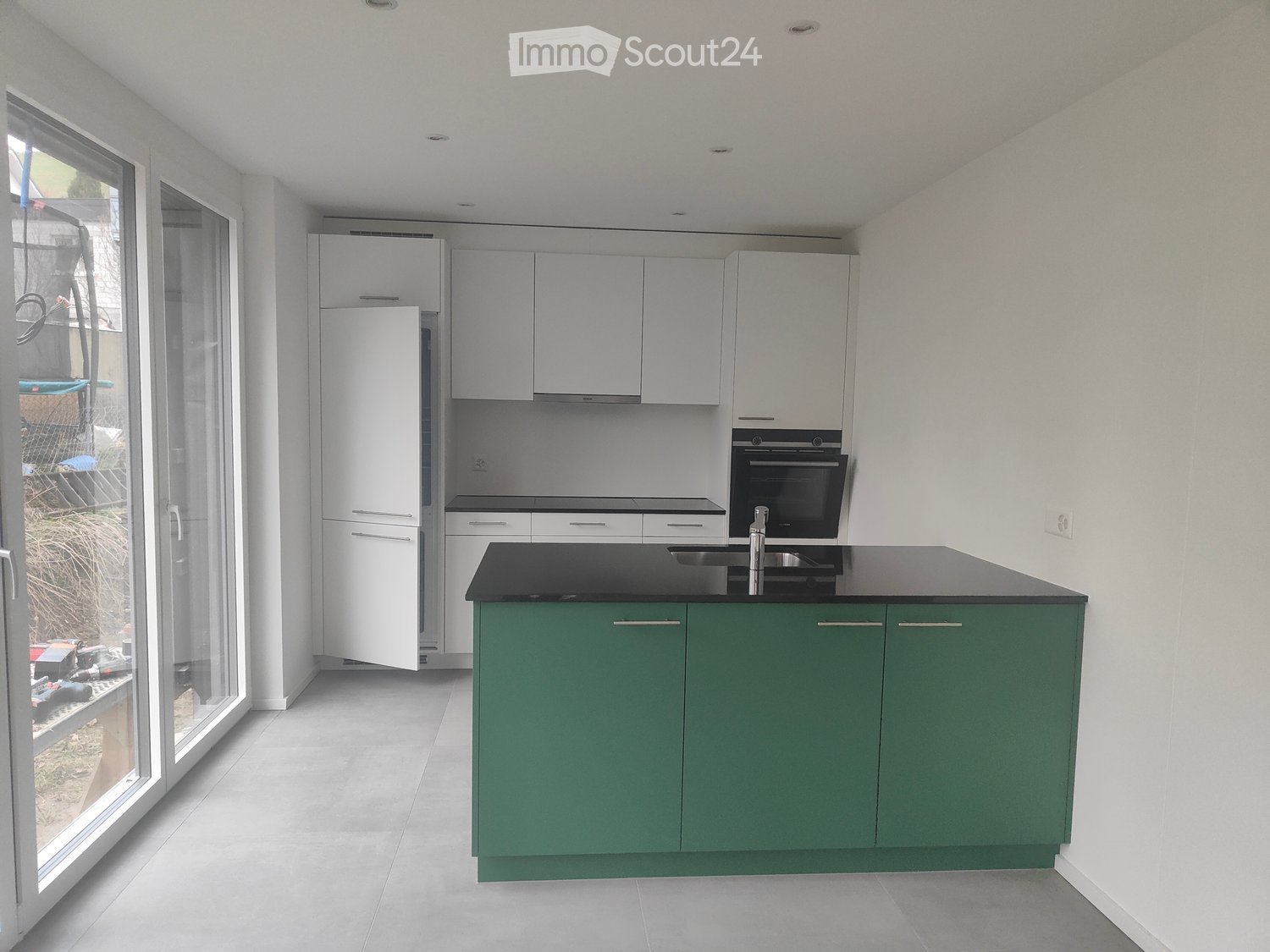 Modern kitchen with green cabinets, black countertops, stainless steel sink and faucet, white appliances, glass door leading to outside.