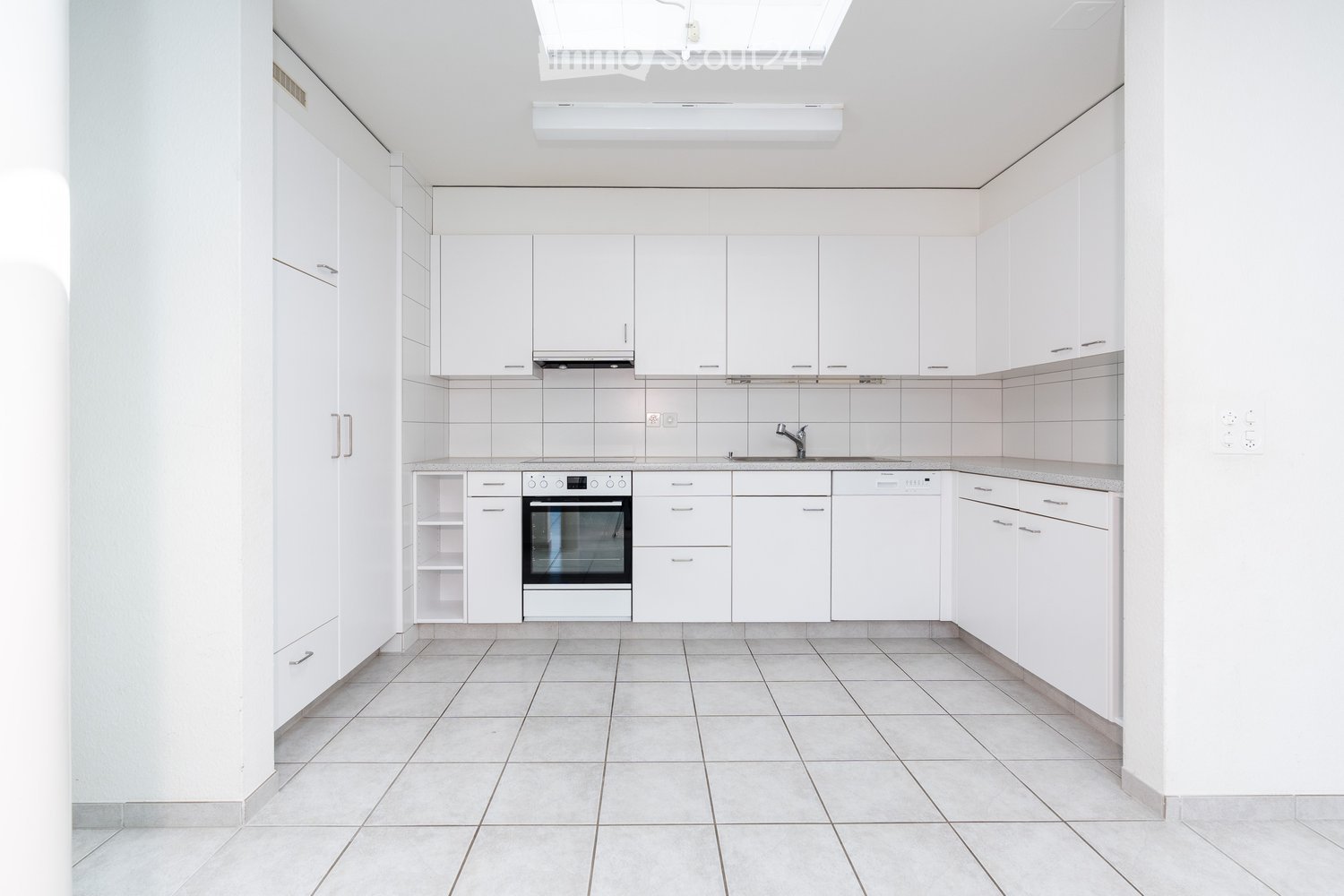 Modern, minimalist kitchen with white cabinets, tiled walls, and a central island featuring an oven and dishwasher. The space is bright with natural light from a skylight.