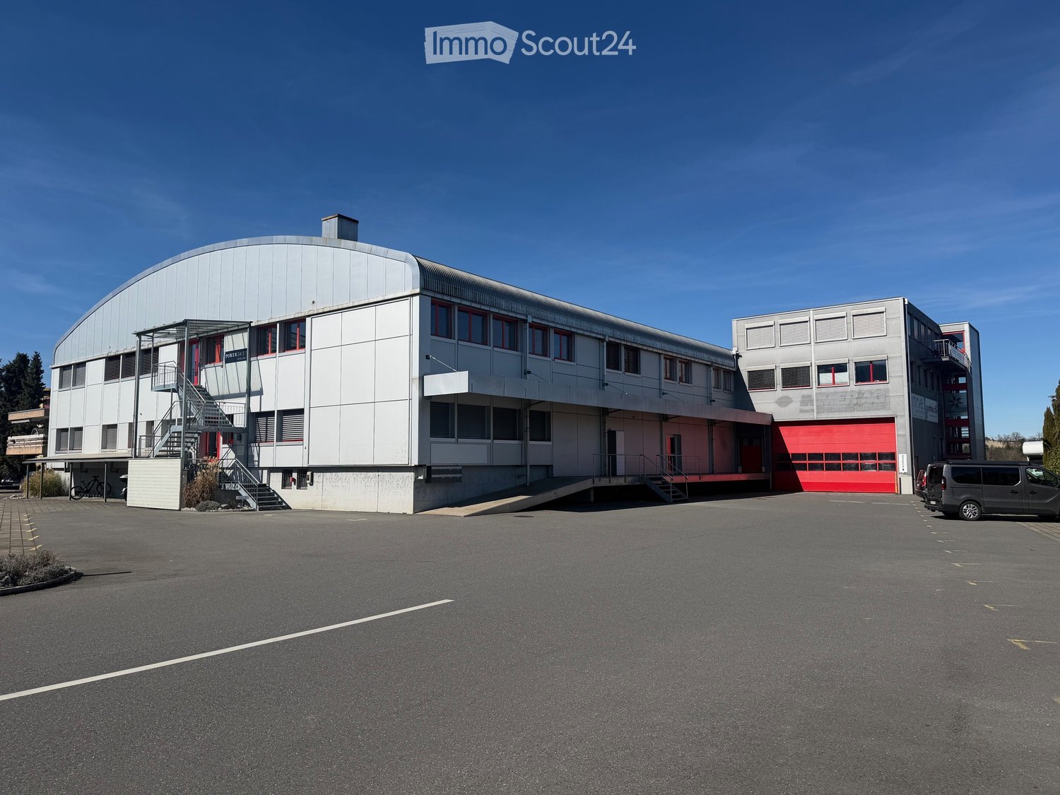 Large industrial building with white walls, red doors, and a large parking area in front
