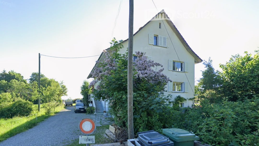 2 story house, white exterior, multiple windows, driveway, stairs, car parked, trees, bushes, trash bins