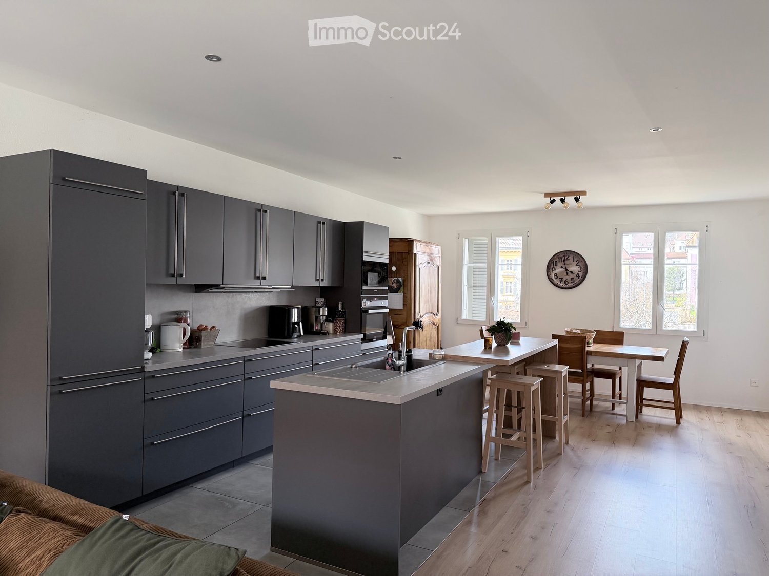 modern kitchen with island, gray cabinets, wooden floor, dining area with table and chairs, large windows, wooden cabinet, clock, potted plants