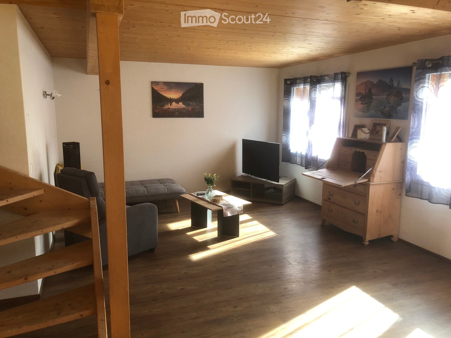 Living room with wooden floor, wooden staircase, couch, coffee table, TV, wooden desk, flower vase, picture frames, curtains