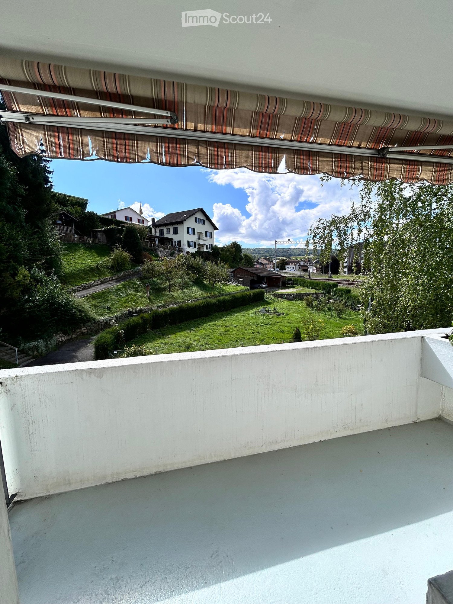 balcony with white railing, white floor, striped retractable canopy, distant view of a green landscape with houses