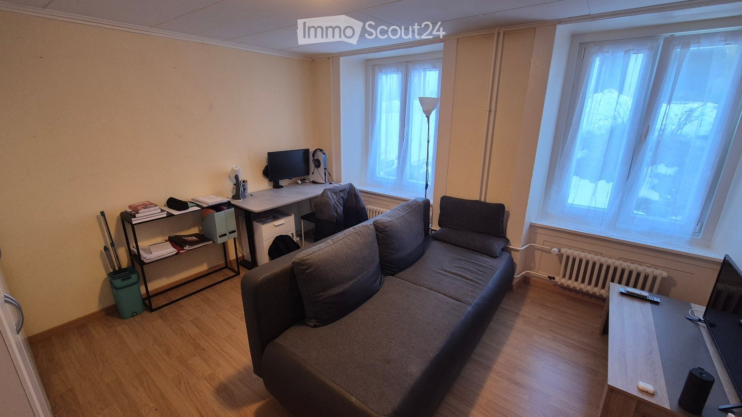 A living room with a couch, a desk, a monitor, a lamp, a radiator, and a television.