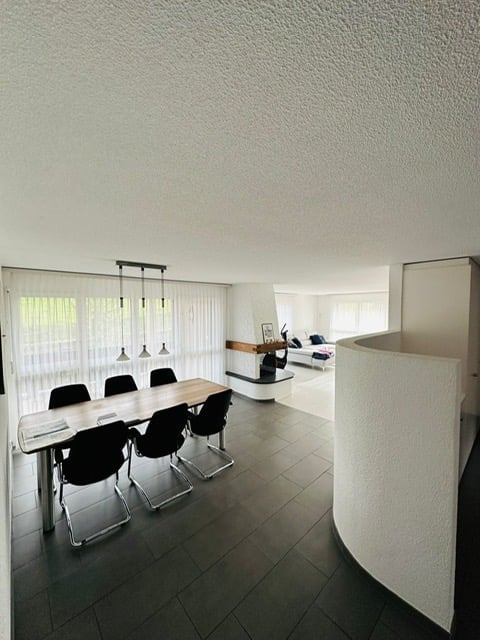 Dining room with a long wooden table, black chairs, white walls, white ceiling, fireplace, white curtains, and a white curved wall.