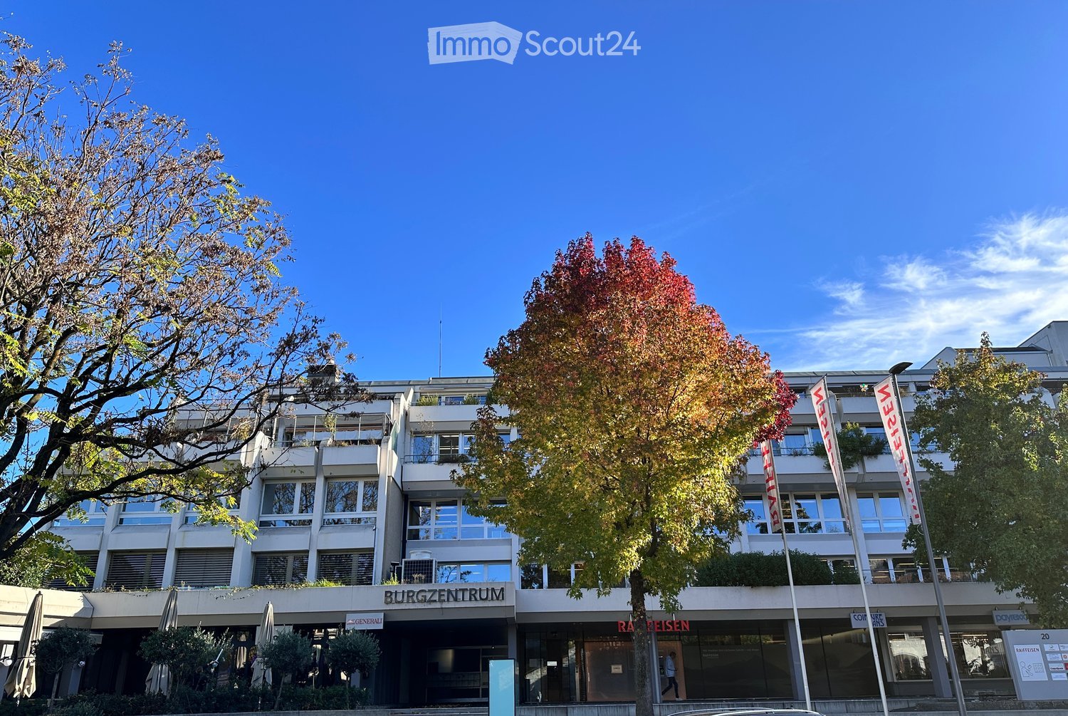 Modern building with glass windows, multiple floors, entrance with sign 'Burgzentrum', flags, trees, and shrubs in front