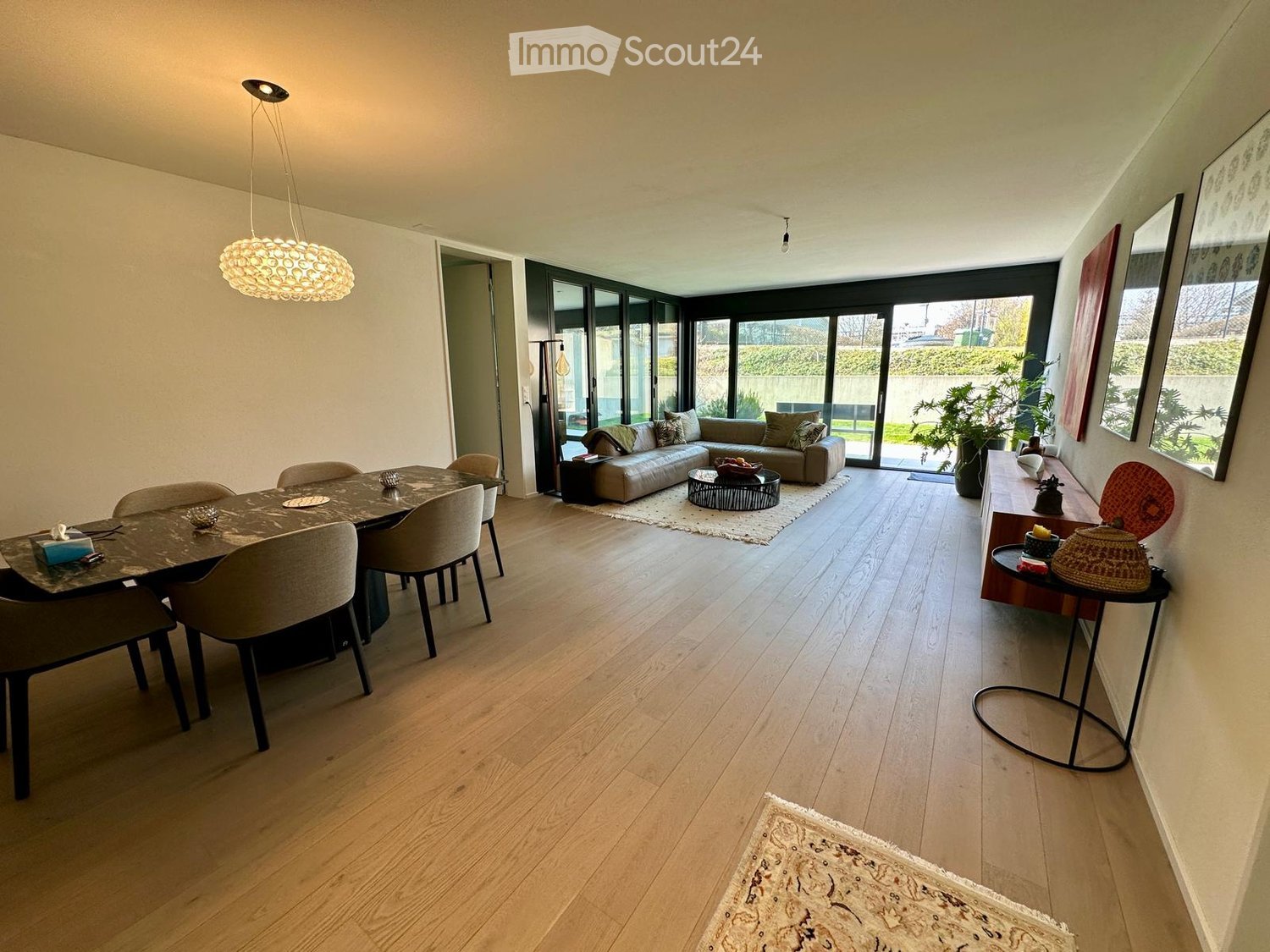 Dining room with wooden floor, large windows, dining table, chairs, couch, coffee table, rug, plants, lamp