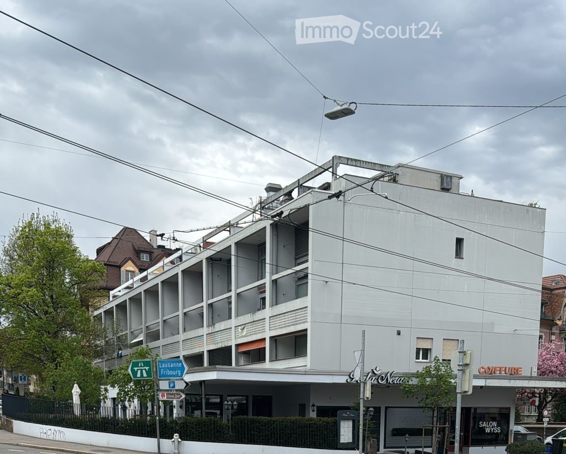 White building with multiple floors, several windows, and a flat roof. Street signs and trees are visible in the foreground.
