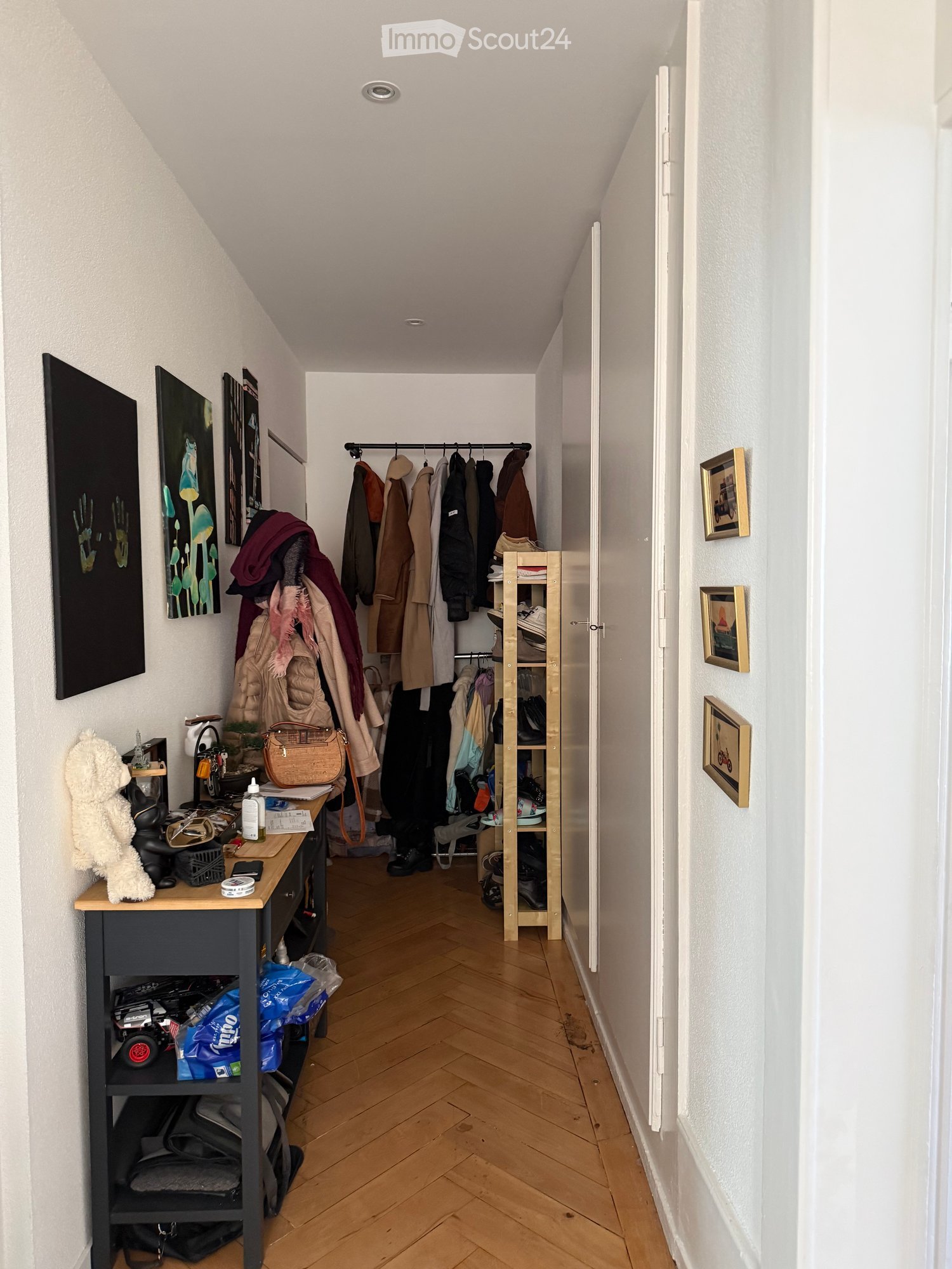 A hallway with wooden floor, white walls, a table with various items, a rack with clothes, and a wooden shoe rack.
