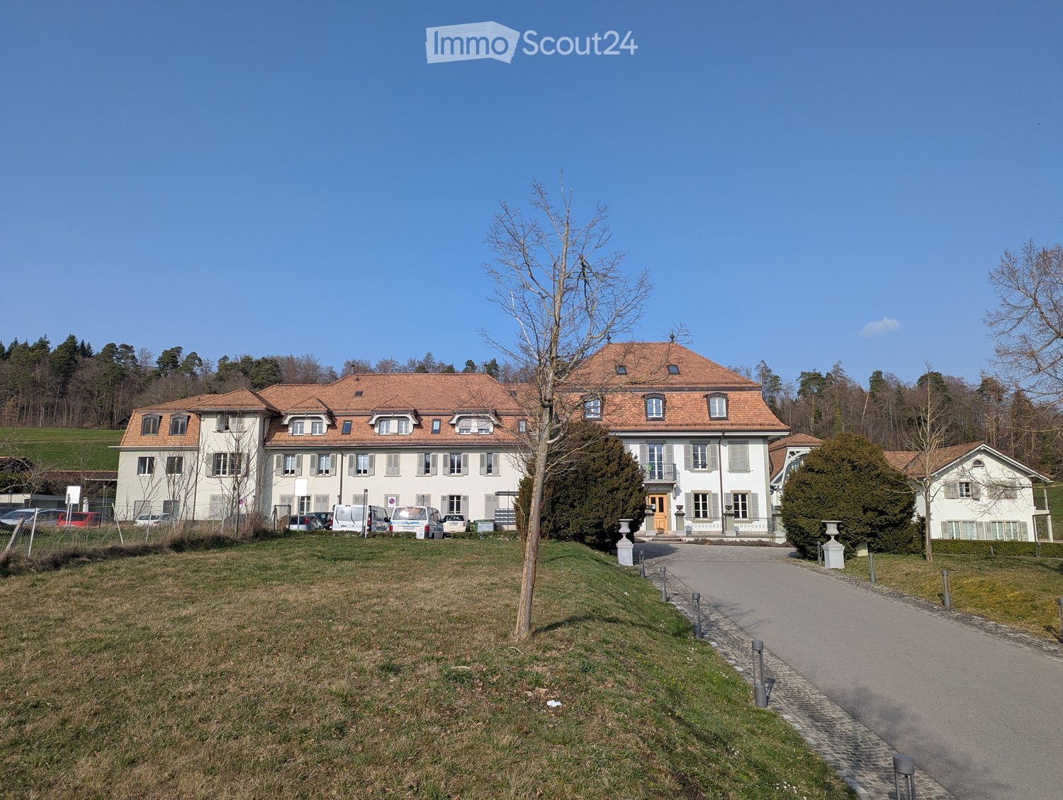 Large white building with brown roof, multiple windows, driveway, cars parked in front, trees, grass, mountains in the background