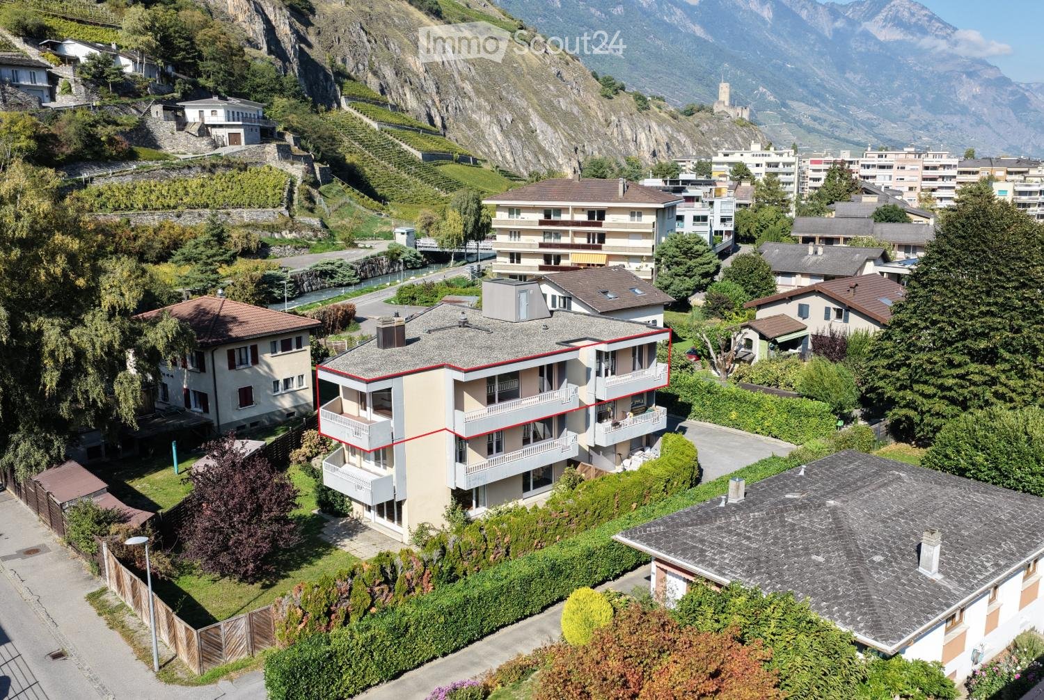 Aerial view of a multi-story building with balconies, surrounded by other buildings and greenery, with a mountain in the background.