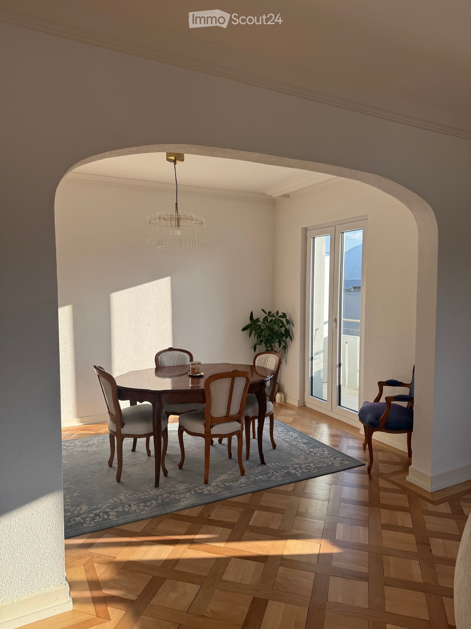 Dining room with wooden table, chairs, chandelier, rug, plant, balcony with sliding glass doors