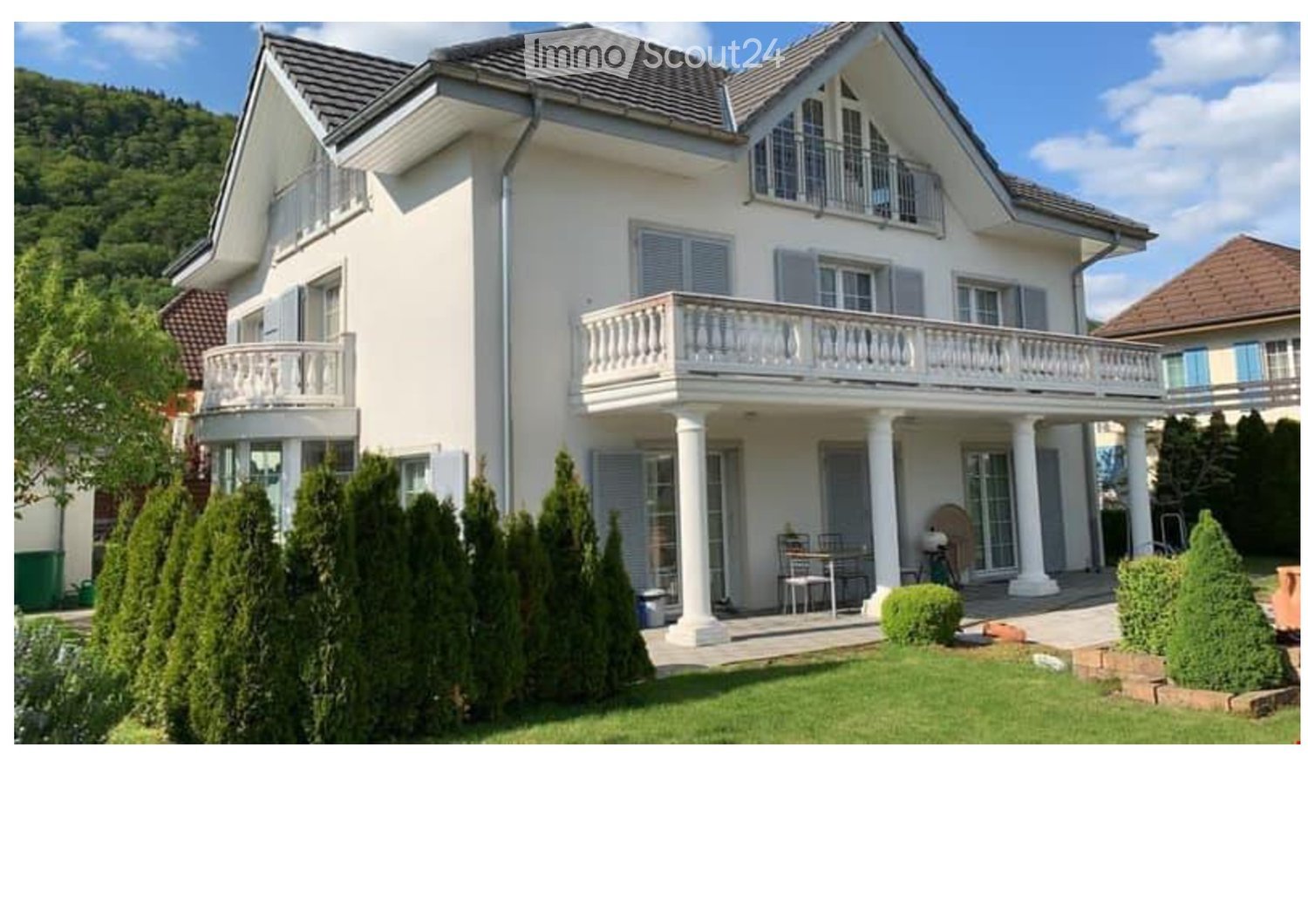 A two-story white house with multiple balconies, large windows, and a well-maintained garden with trimmed trees and shrubs.