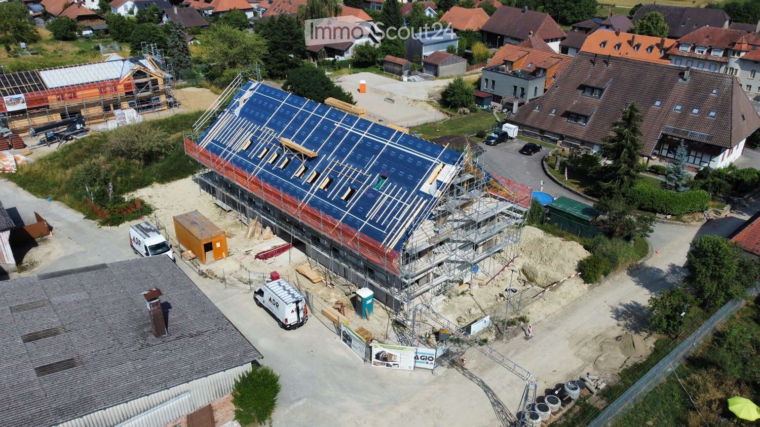 Aerial view of a residential area with a house under construction, surrounded by other houses, trees, and parked vehicles.
