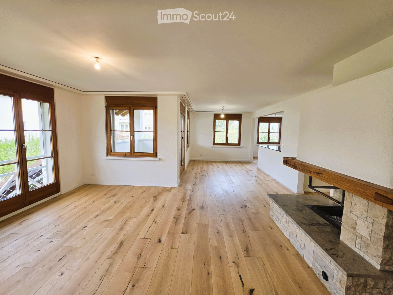 Empty room with hardwood floors, white walls, large windows, and a fireplace with a stone mantle.