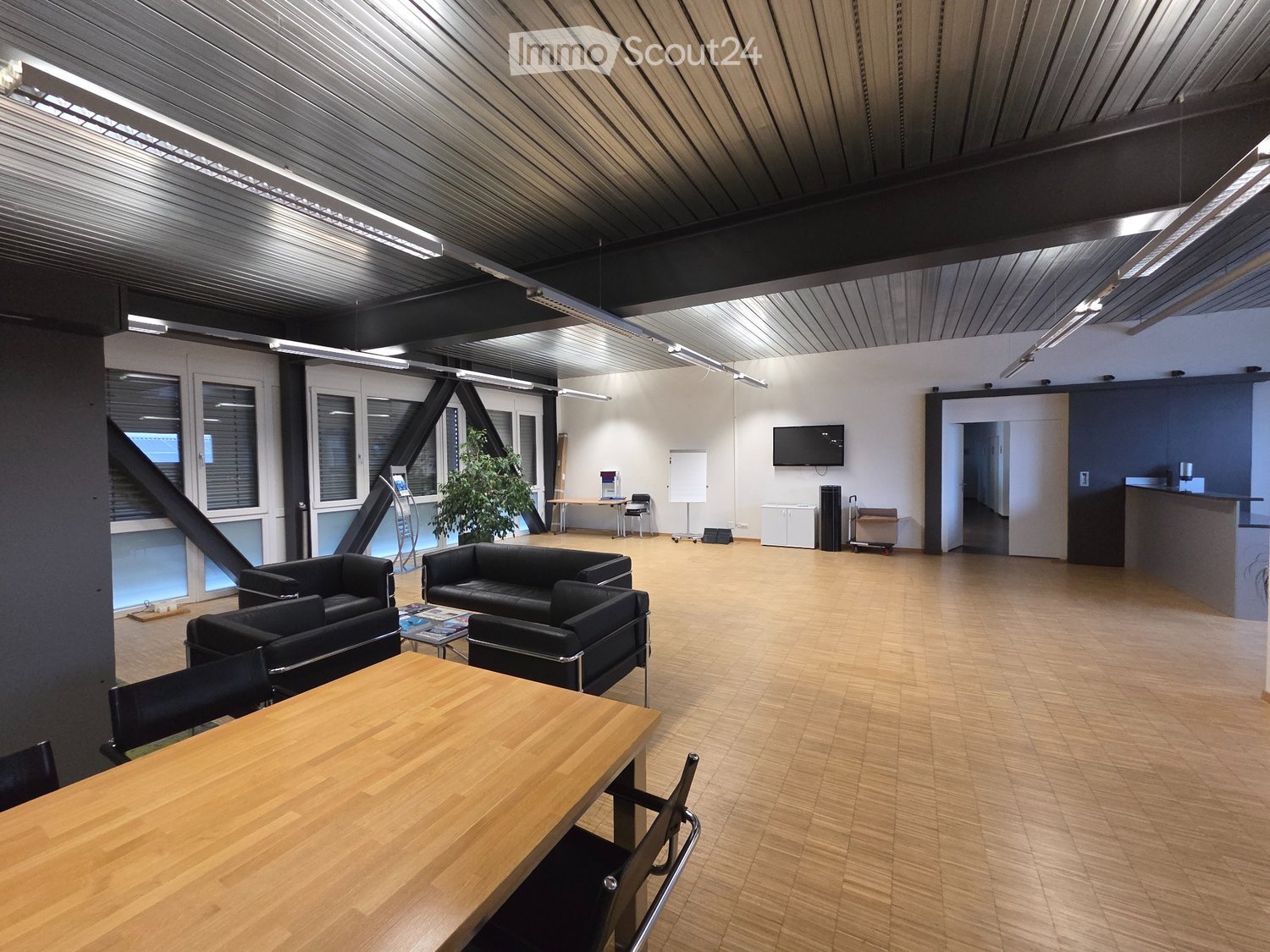 large open room, wooden floor, black couches, wooden table, mounted TV, white walls, ceiling lights