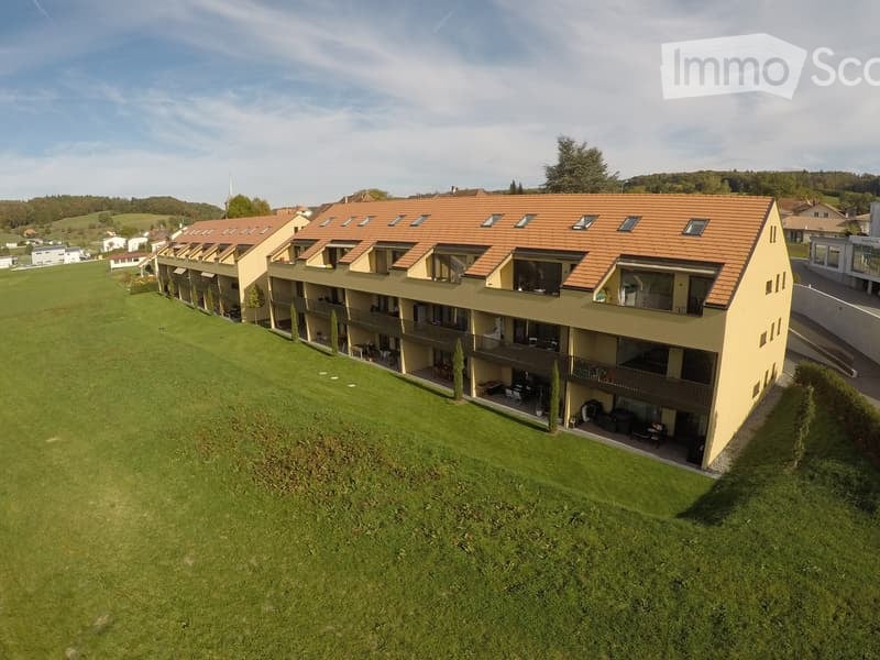 Apartment building with multiple floors, balconies, and a brown roof, set in a large green field