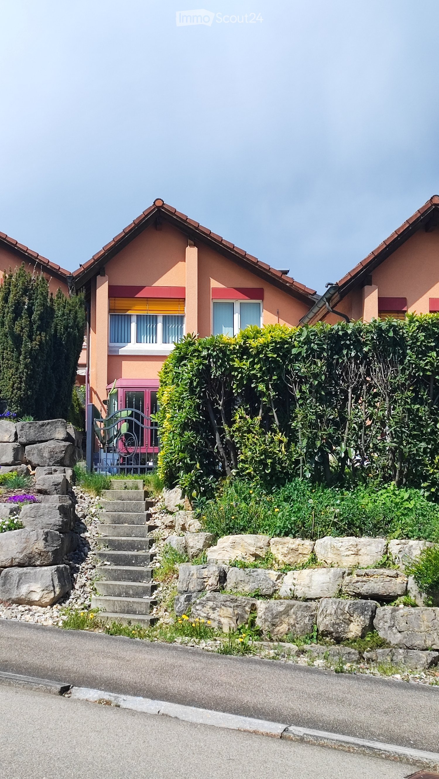 Two-story house with peach-colored walls, red roof, and white-framed windows. A stone staircase leads up to the entrance, surrounded by greenery and rocks.