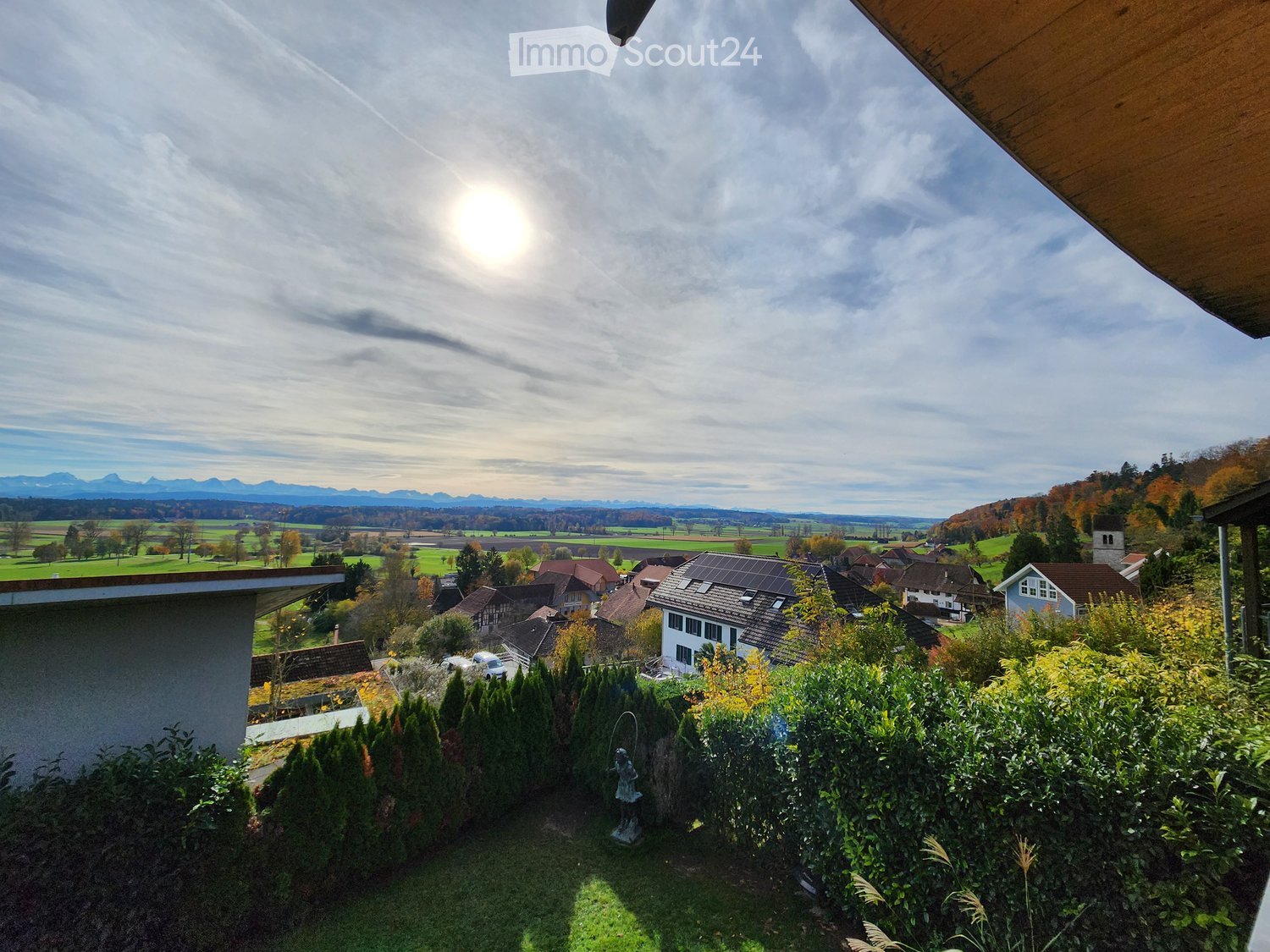 A scenic view from a balcony or terrace overlooking a village with houses, green fields, and distant mountains under a partly cloudy sky.