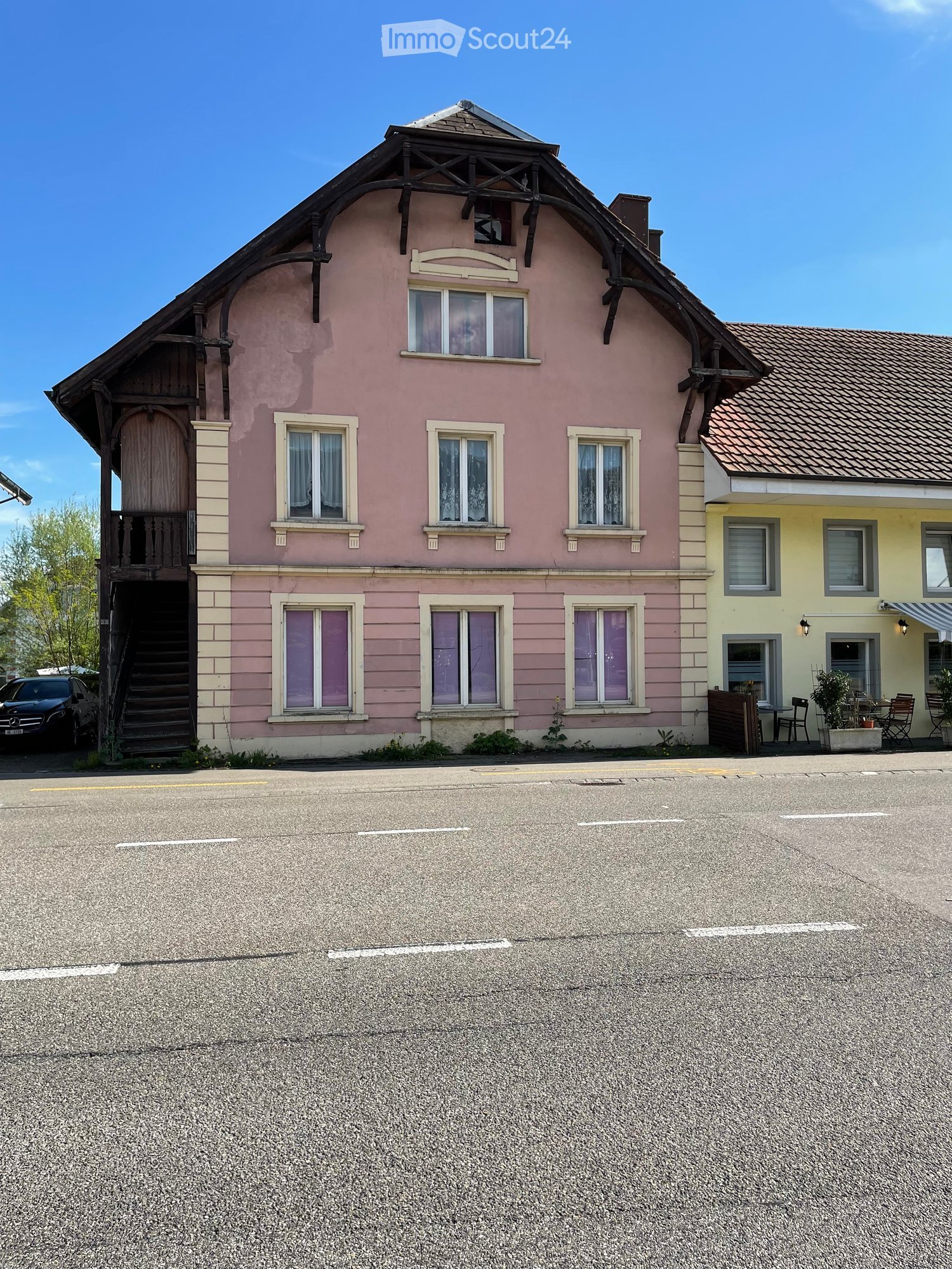 2 story house, pink exterior, wooden balcony, multiple windows, stairs leading to the entrance, parked car in front