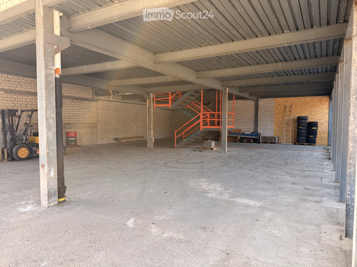 Empty warehouse with metal beams, brick walls, concrete floor, orange staircase, forklift, tires, ladder