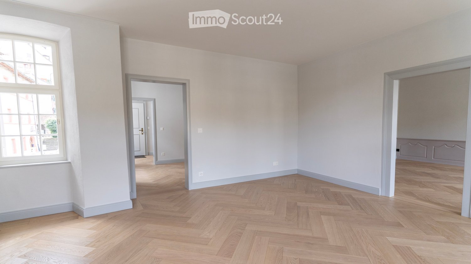 Empty room with white walls, wooden parquet floor, large window, two doors leading to other rooms