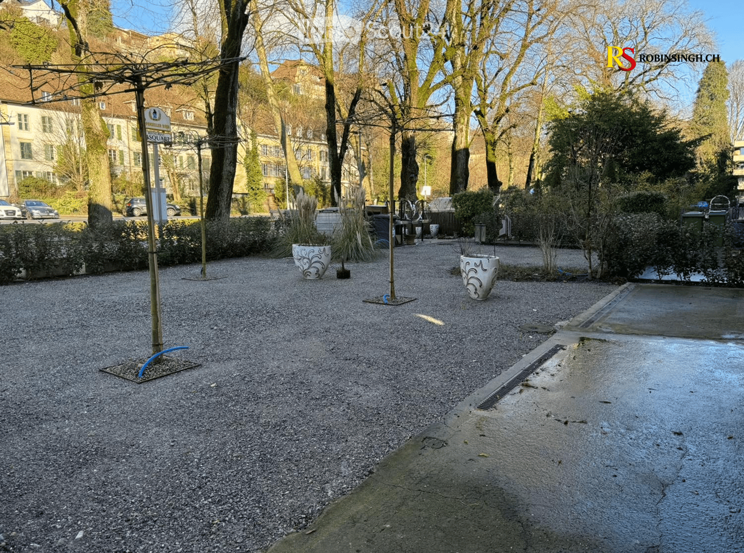 gravel ground, two large white vases, metal poles, bushes, trees, buildings in the background