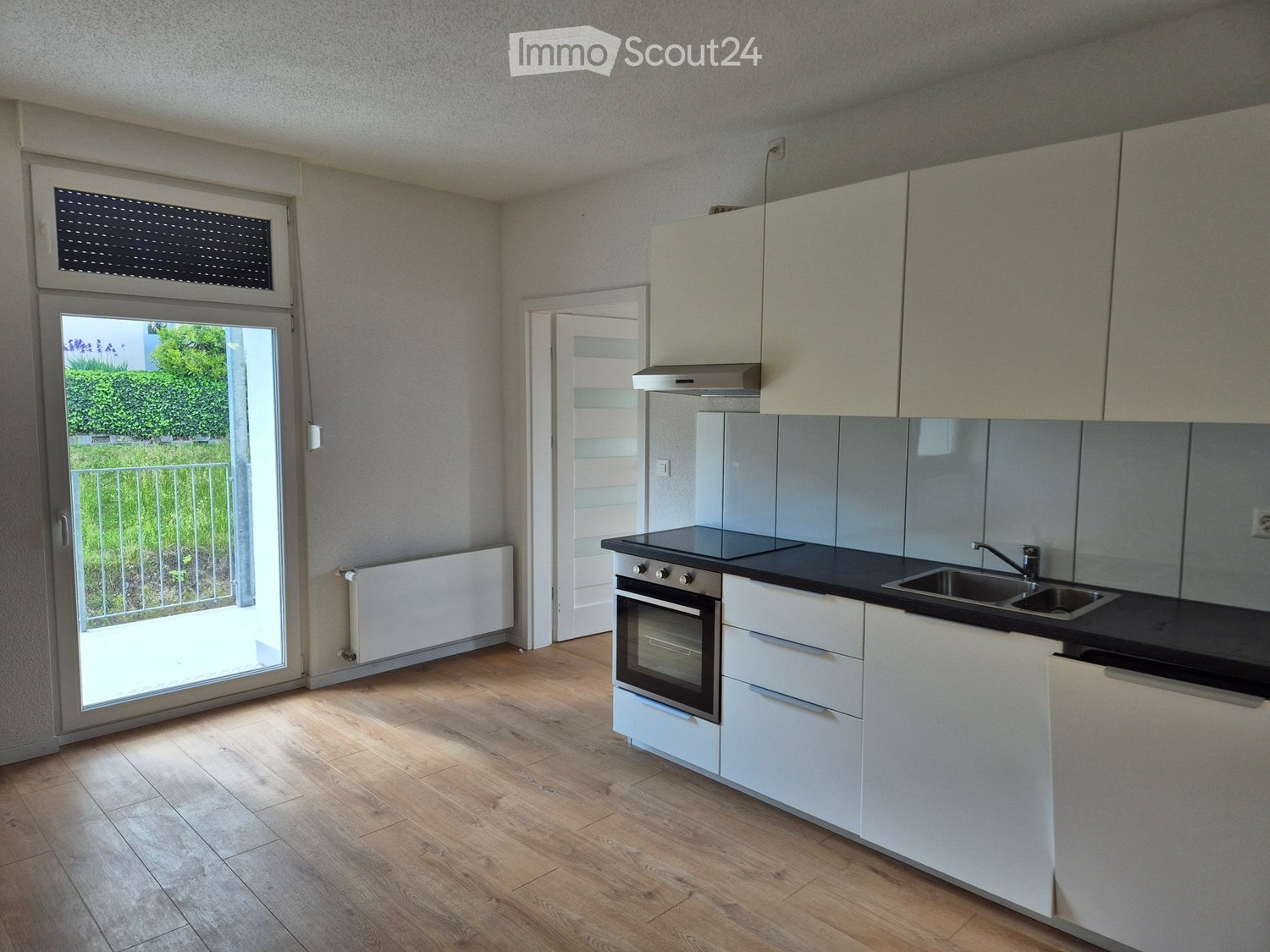 Modern kitchen with white cabinets, black countertops, an oven, a sink, and a range hood. The space features wooden flooring and large windows with blinds, offering a bright and airy atmosphere.