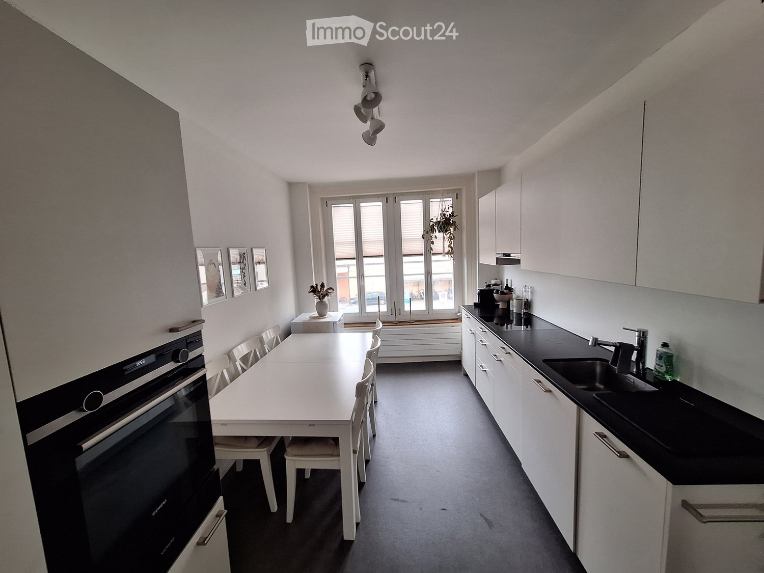 modern kitchen with white cabinets, black countertops, white dining table, white chairs, white refrigerator, oven, sink, faucet, bottles, mounted lights, windows, potted plant, picture frames
