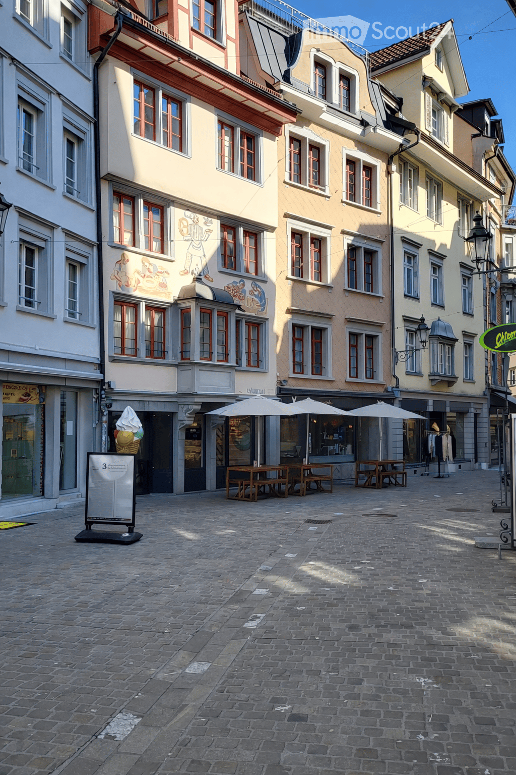 Cobblestone street, multiple buildings, outdoor seating, street lamps, storefronts, mural