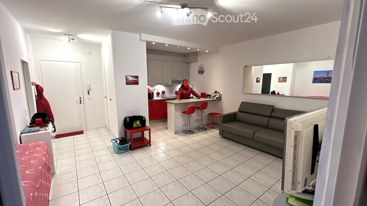 Open plan living room and kitchen, white tiles, gray couch, red stools, kitchen island, mirror, white cabinets, red counter, person in red jacket