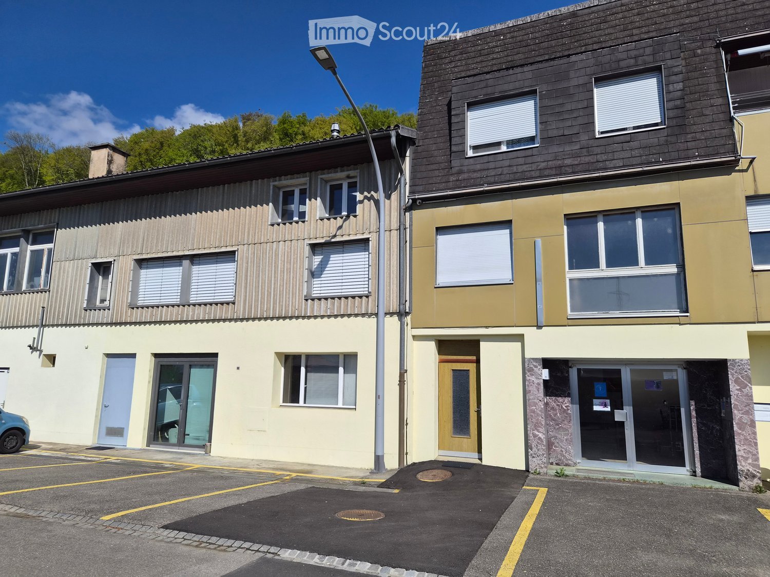 Three-story building with multiple windows, glass doors, and a parking area in front.
