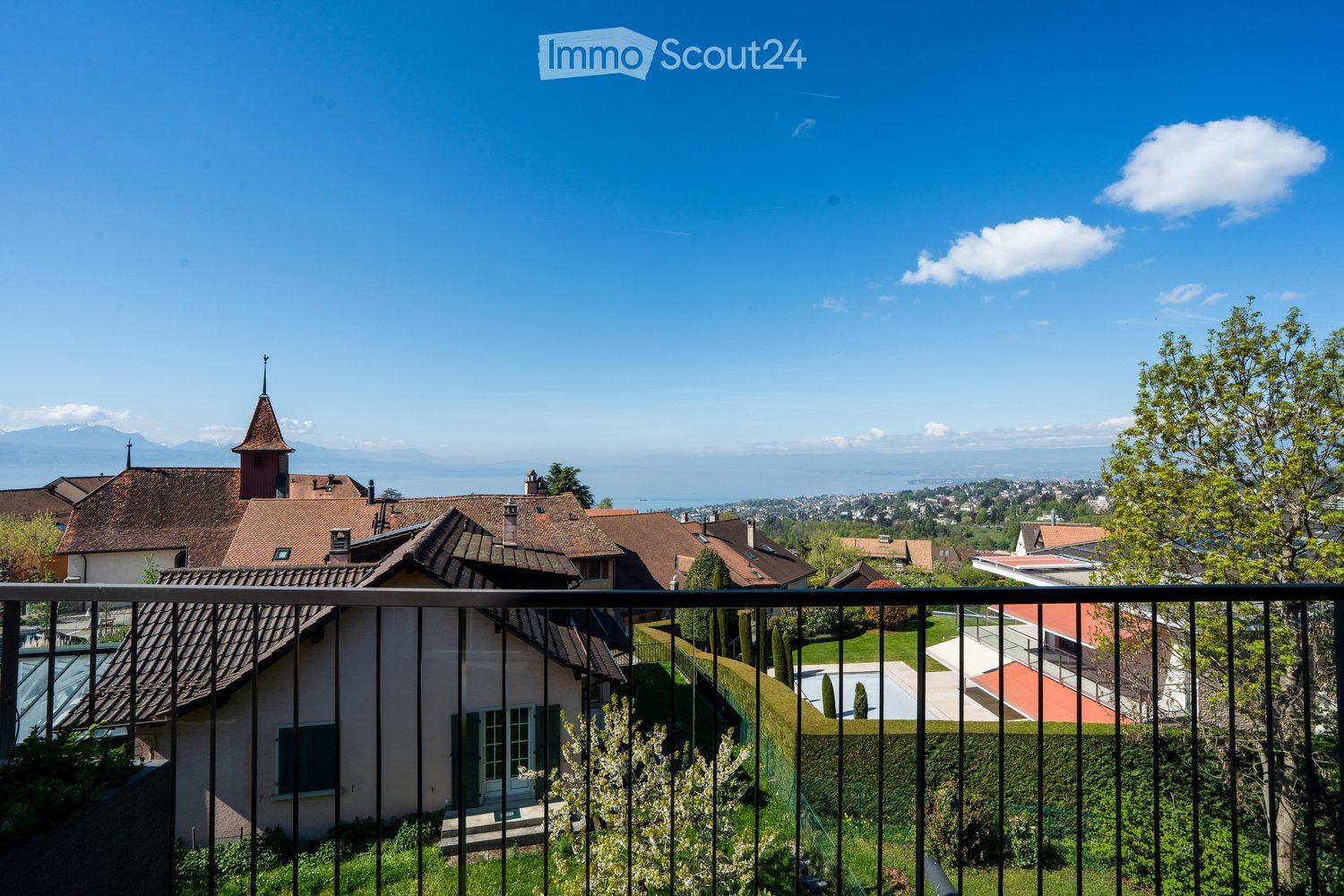House with brown roof, white windows, surrounded by greenery, balcony with black railings, distant view of mountains and city