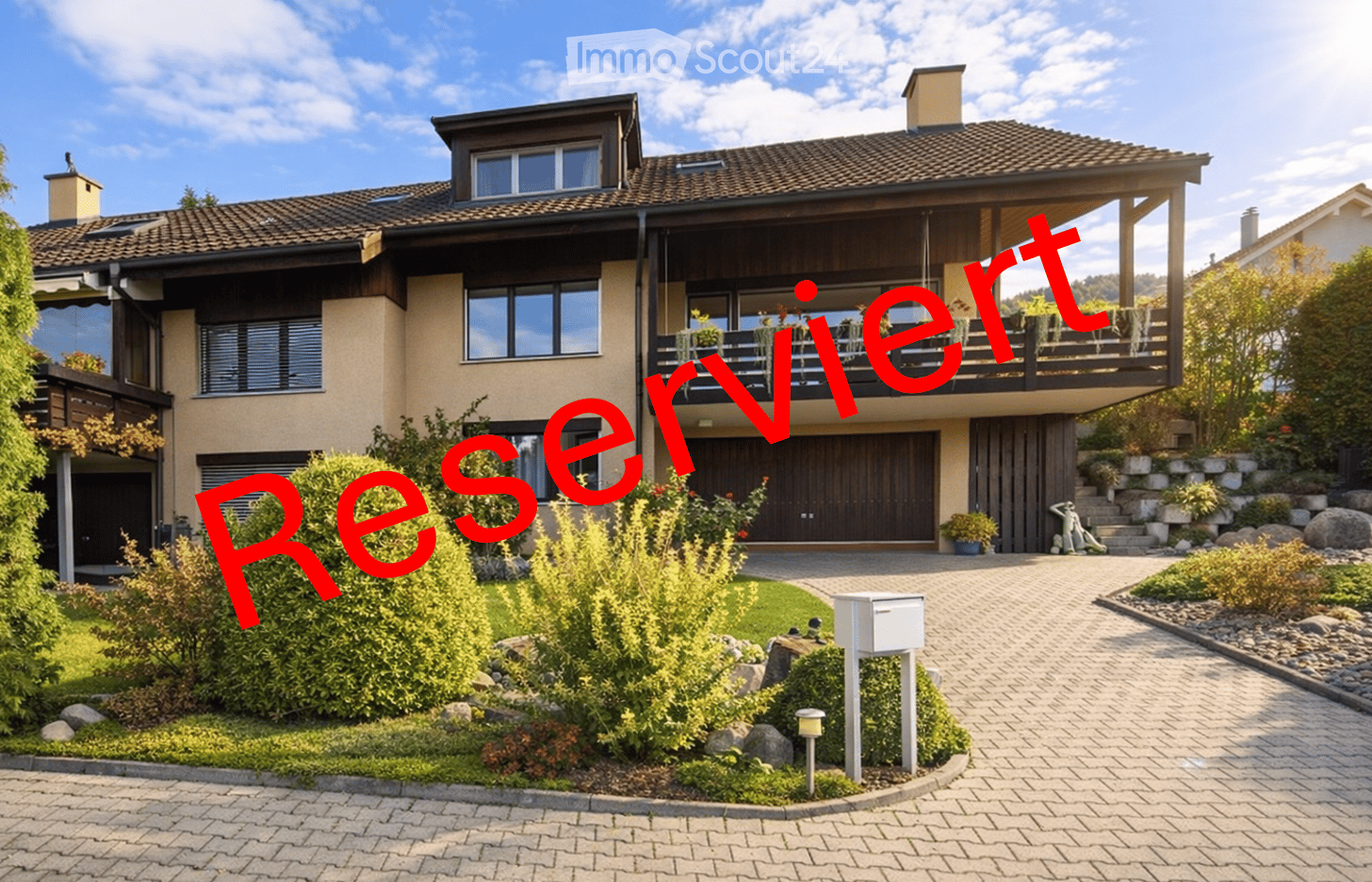 Two-story house with a tiled roof, large windows, and a balcony on the upper level. The house features a well-maintained garden with shrubs and a paved driveway.
